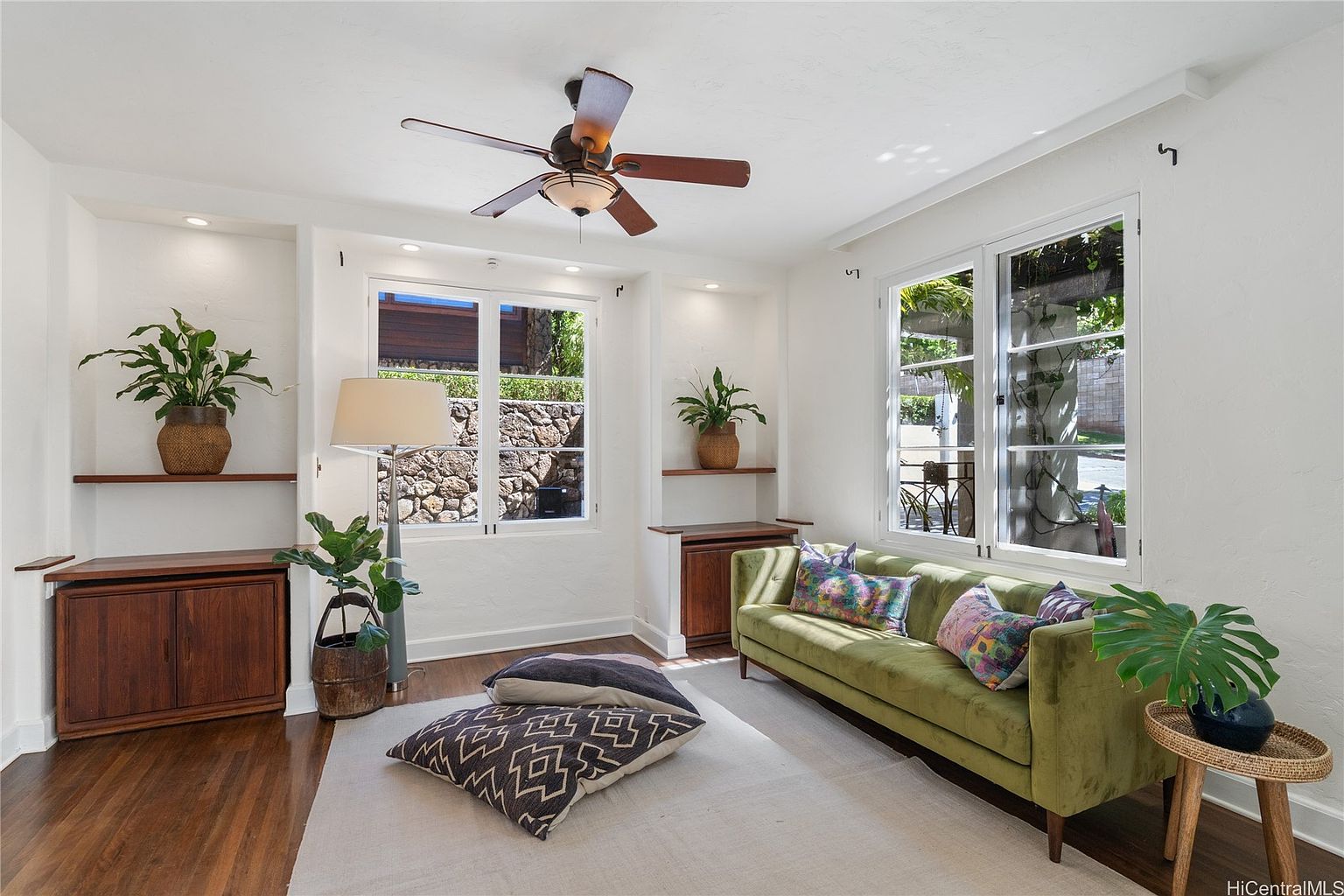 This is an interior shot of a living room featuring a green sofa with colorful pillows, a light-colored rug, and hardwood floors. The room has white walls with built-in shelving and cabinets, and natural light streams in through two windows, creating a bright and inviting atmosphere. A ceiling fan is visible, and plants are placed throughout the room, adding a touch of nature.