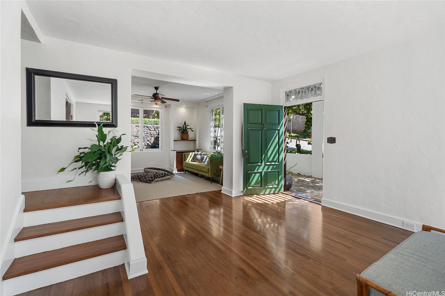 This interior shot showcases a bright and inviting living room with hardwood floors and white walls. A green front door is slightly ajar, leading to the outside. The room features a staircase with a large mirror, and a glimpse into a cozy seating area with a green sofa and a ceiling fan, creating a warm and welcoming atmosphere.