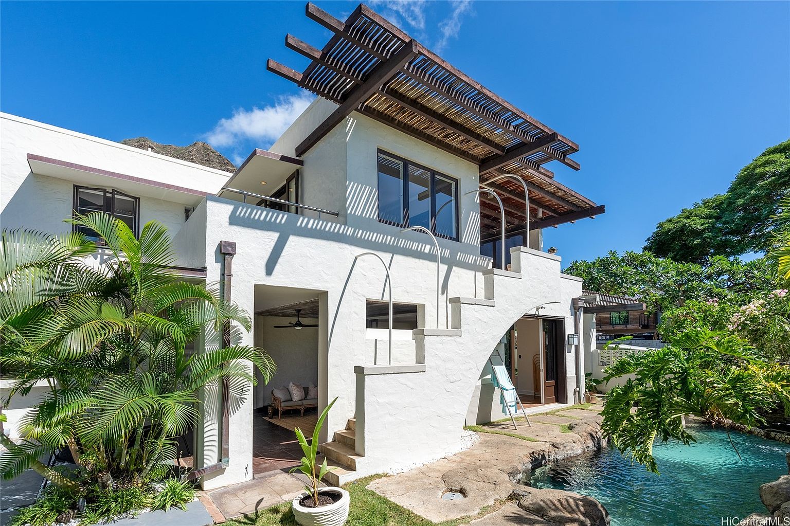 This image showcases the rear exterior of a modern home, highlighting its unique architectural design with a pergola-covered upper level and an integrated pool area. The white stucco facade contrasts with the wooden pergola, while lush tropical landscaping surrounds the property, creating a private and serene backyard oasis. The pool's naturalistic design blends seamlessly with the surrounding rock features.