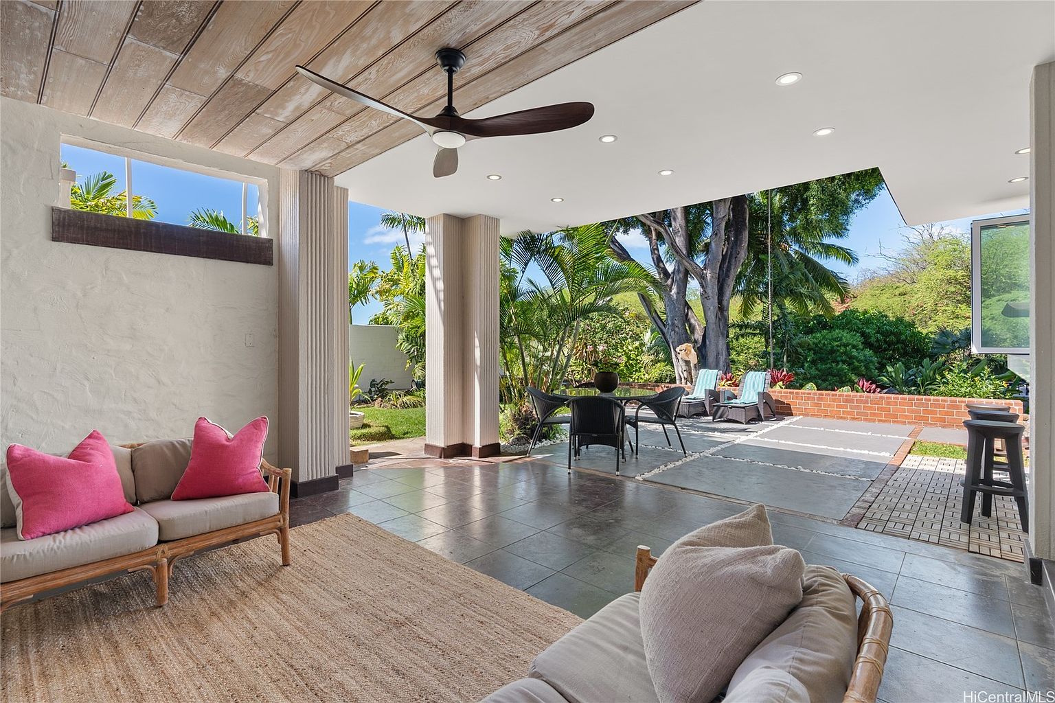 This image showcases a covered outdoor patio area, seamlessly blending indoor and outdoor living. The space features comfortable seating arrangements, including a sofa with pink pillows and a rattan chair, complemented by a dining set. The patio is adorned with a ceiling fan and overlooks a lush garden, creating a serene and inviting atmosphere.