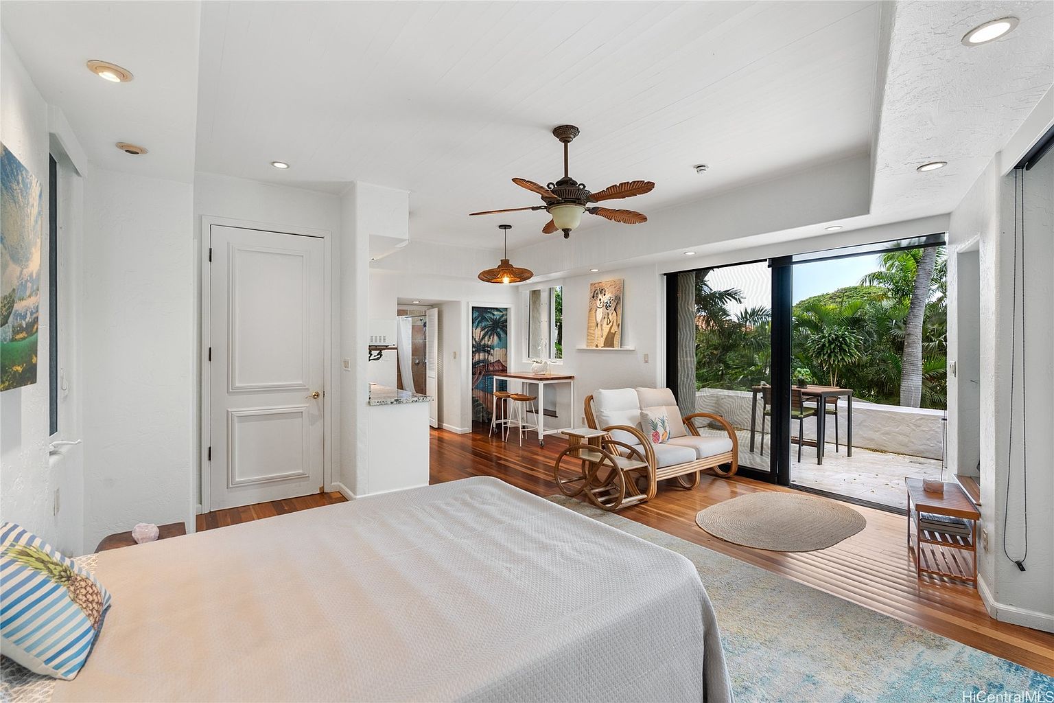 This is a bright and airy primary bedroom featuring hardwood floors, white walls, and a ceiling fan. The room includes a bed with a light-colored comforter, a seating area with a rattan loveseat, and a sliding glass door leading to a patio. The overall style is relaxed and tropical, creating a serene and inviting atmosphere.