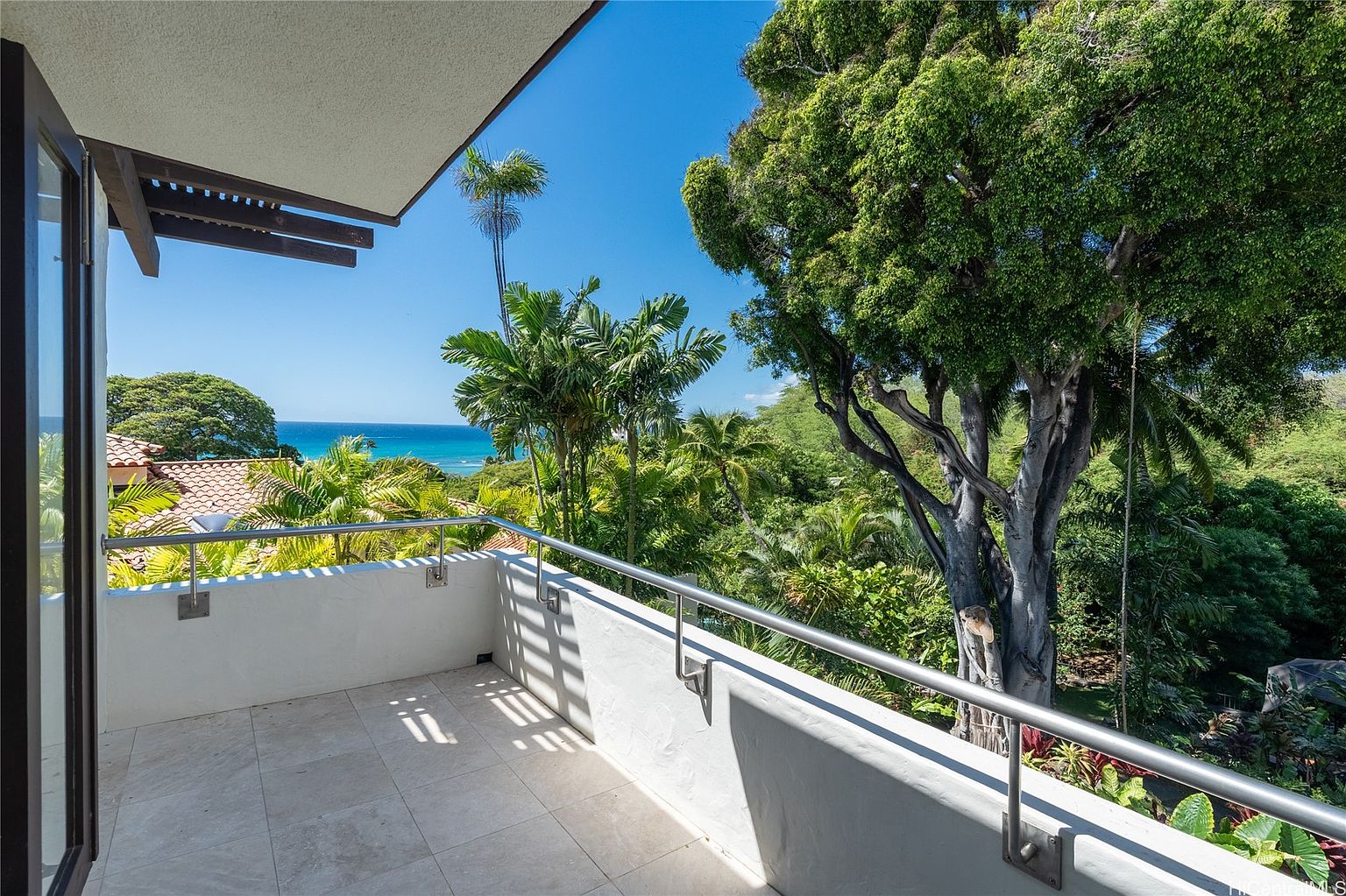 This image showcases a balcony with a view of the ocean and lush greenery. The balcony features a white railing with a stainless steel handrail and a tiled floor. The view includes palm trees, other tropical plants, and a glimpse of the ocean, creating a serene and inviting atmosphere.