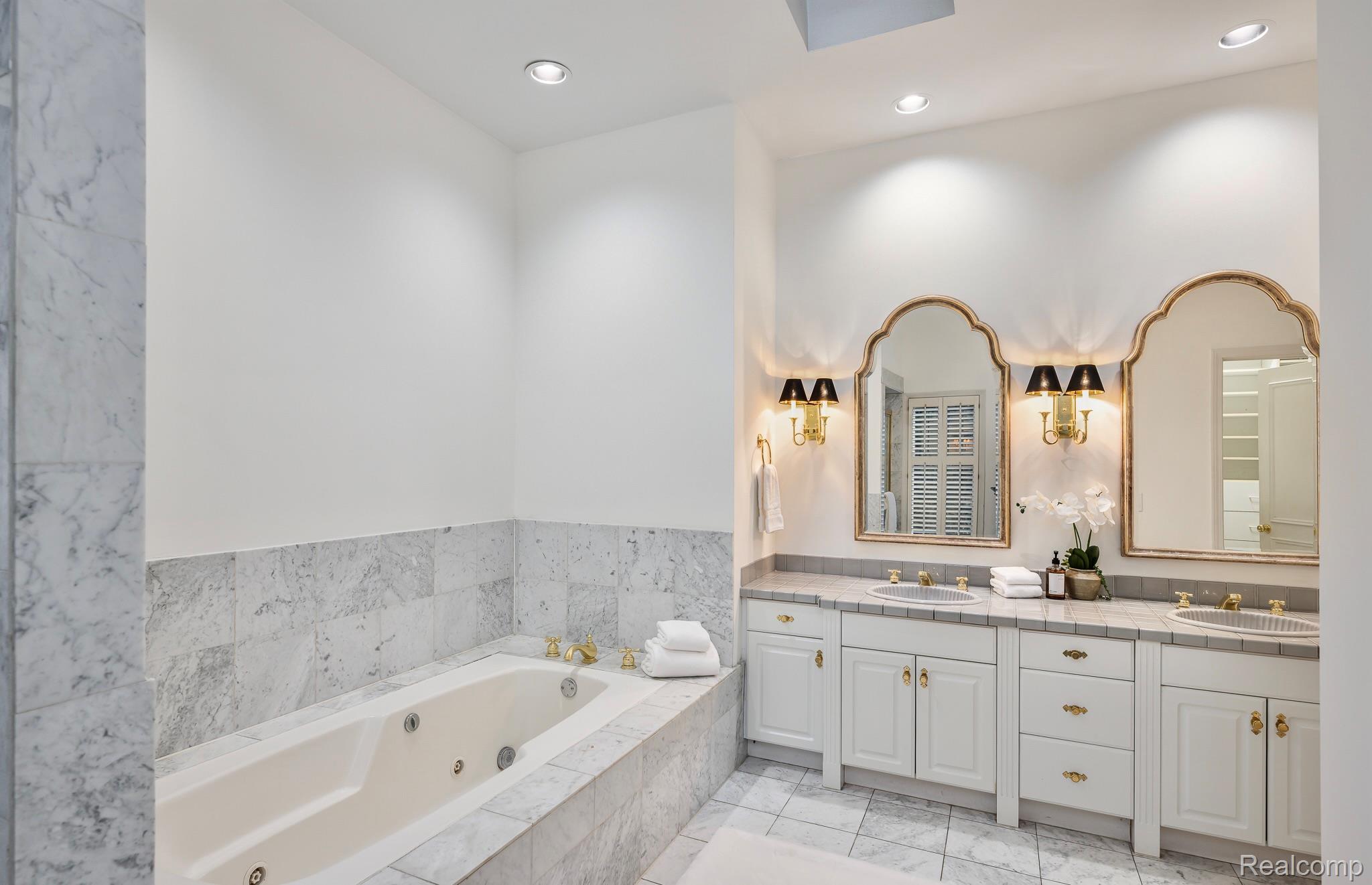 This is a luxurious primary bathroom featuring a large soaking tub with marble surround and a double vanity with ornate gold-framed mirrors. The cabinetry is white with gold hardware, and the countertops are a light gray tile. The room is well-lit with recessed lighting and sconces, creating a bright and elegant space.