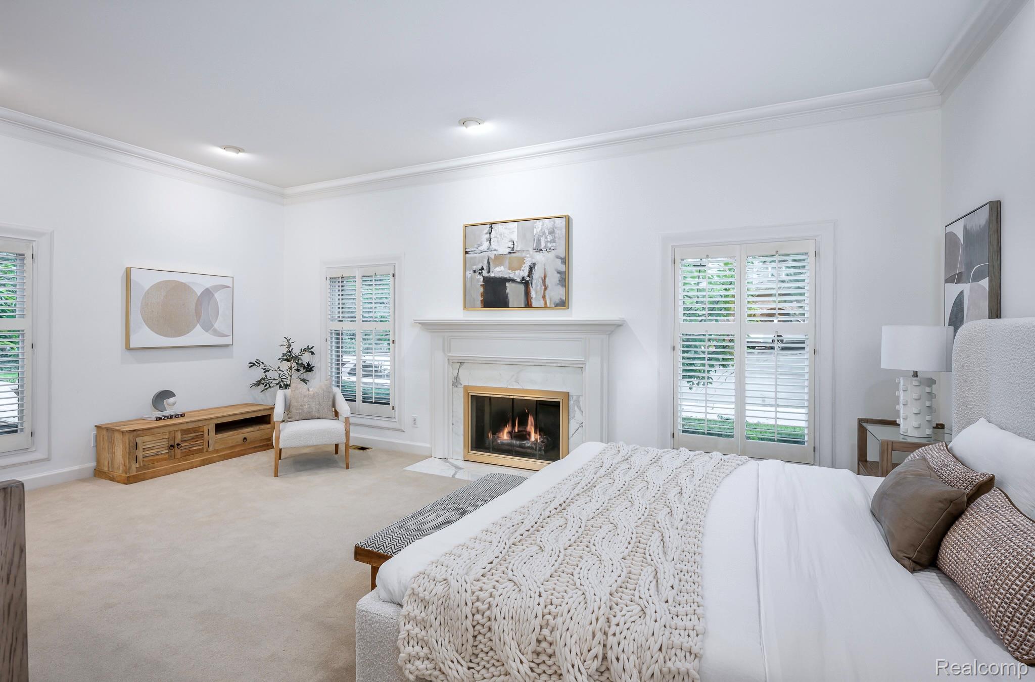 This is a bright and spacious primary bedroom featuring a fireplace, neutral color palette, and ample natural light from the shuttered windows. The room is furnished with a large bed, a wooden media console, and a comfortable armchair, creating a relaxing and inviting atmosphere. The overall impression is one of comfort and luxury.