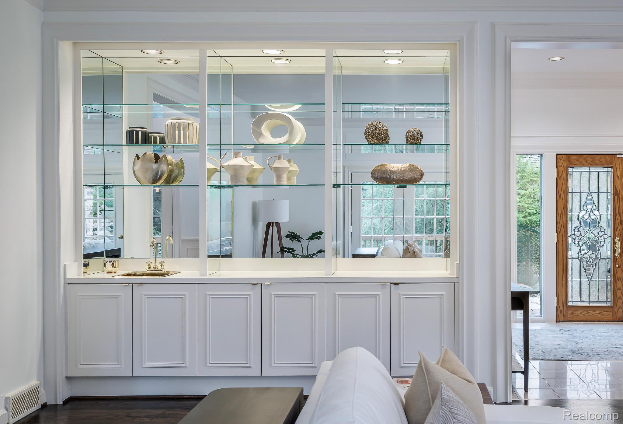 This interior shot showcases a built-in display cabinet filled with decorative items, positioned between the living room and an entryway. The cabinet features glass shelves, mirrored backing, and white cabinetry below, creating a sophisticated focal point. The open doorway provides a glimpse into another room, enhancing the sense of spaciousness.