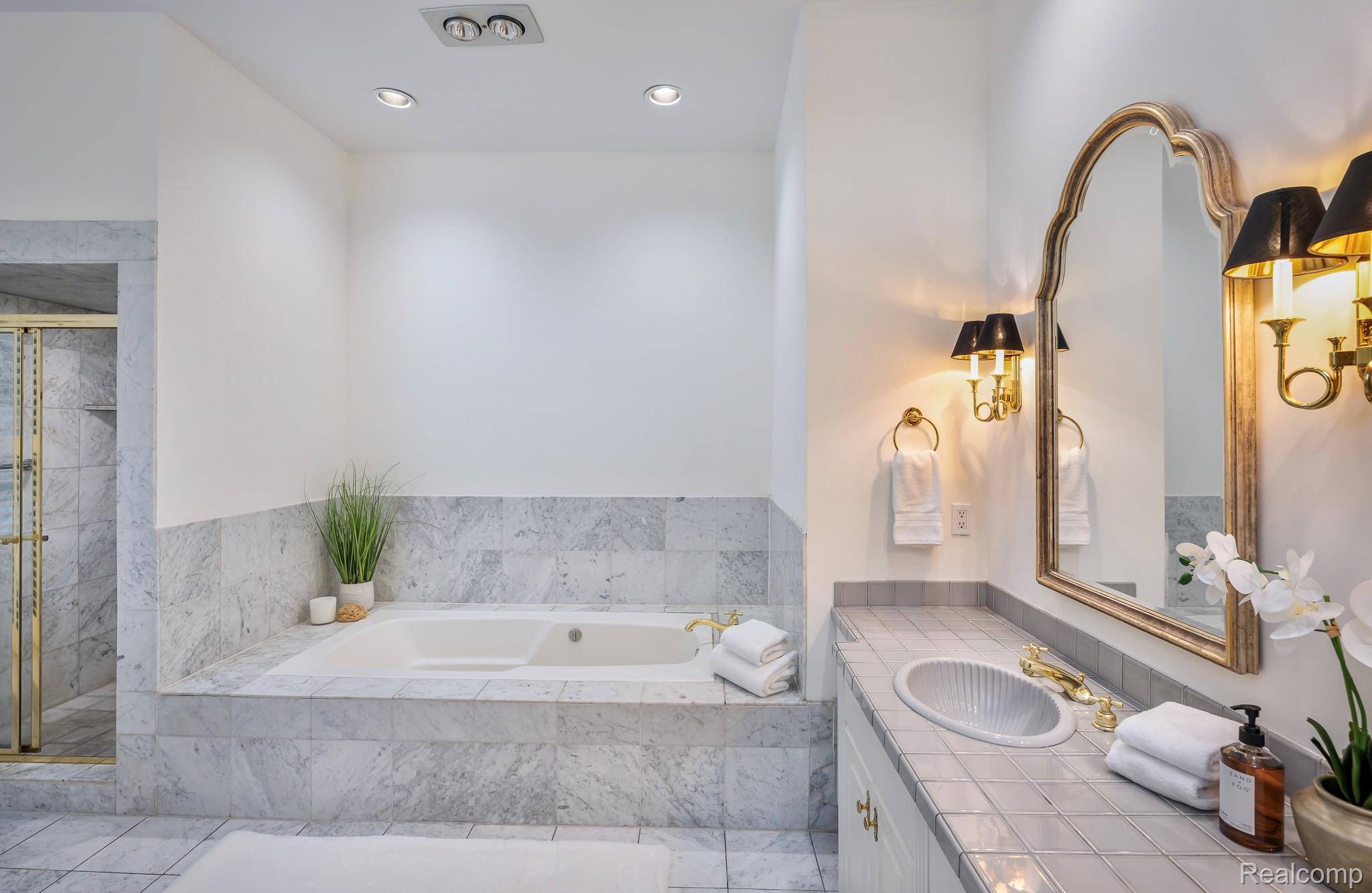 This is a primary bathroom featuring a marble-tiled bathtub with a white interior, complemented by a marble surround. A vanity with a light gray tiled countertop and a gold-framed mirror with sconces adds a touch of elegance. The overall impression is clean, bright, and luxurious.