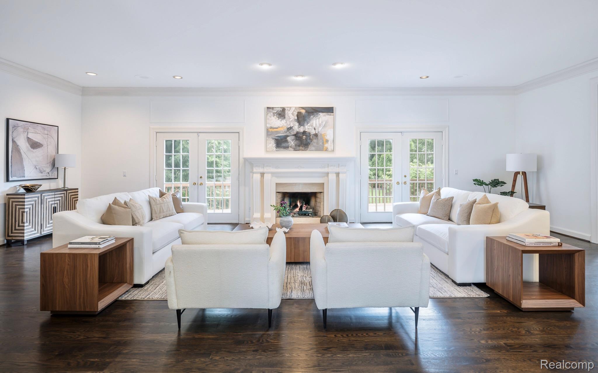 This is a bright and spacious living room featuring two white sofas, two white armchairs, and a wooden coffee table. The room has a fireplace with an abstract painting above it, and double doors leading to the outside. The dark hardwood floors and wooden side tables add warmth to the space, creating an inviting and elegant atmosphere.