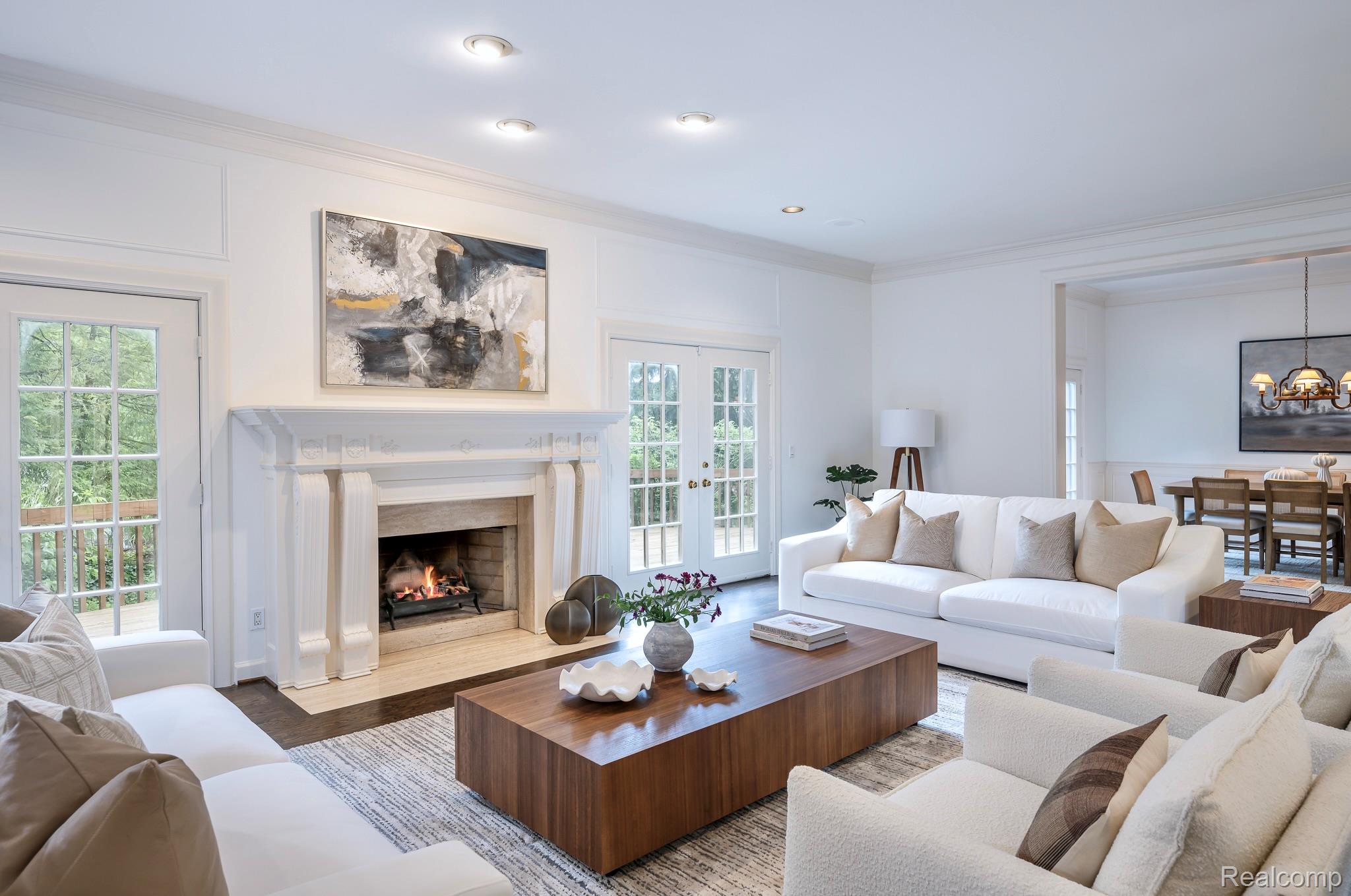 This is a bright and airy living room featuring white walls, a fireplace, and a large wooden coffee table. The room is furnished with white sofas and armchairs, creating a comfortable and inviting space. The open layout connects to the dining area, enhancing the sense of spaciousness.