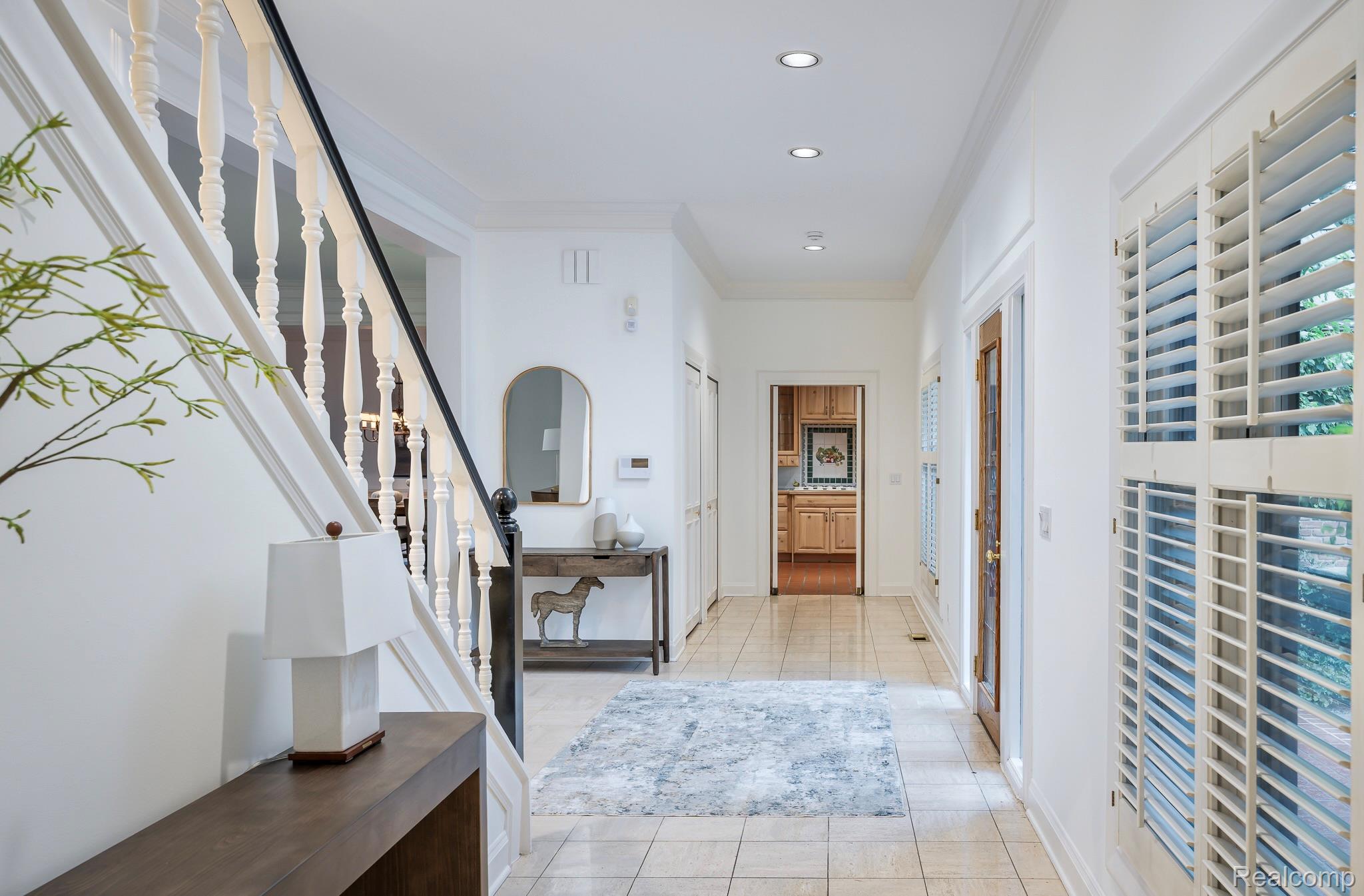 The image showcases a bright and inviting hallway with a staircase. The hallway features light-colored tile flooring, white walls, and recessed lighting. A wooden console table with a lamp sits near the staircase, and a decorative mirror and artwork adorn the walls, creating a welcoming and elegant entrance.