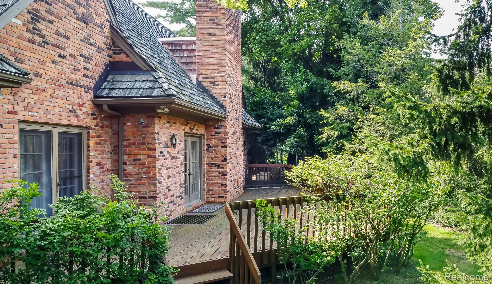The image showcases a charming brick house with a wooden deck and lush greenery. The deck features wooden railings and steps, leading to a well-maintained yard. The brick exterior and the surrounding trees create a cozy and inviting atmosphere, perfect for outdoor relaxation and entertainment.