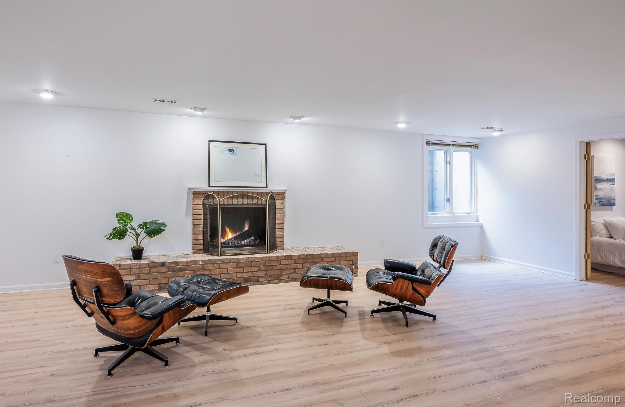 This is an interior shot of a living room featuring a brick fireplace with a decorative screen, flanked by two modern lounge chairs and an ottoman. The room has light wood flooring, white walls, and recessed lighting, creating a bright and inviting atmosphere. A window provides natural light, and a doorway leads to another room, suggesting an open floor plan.
