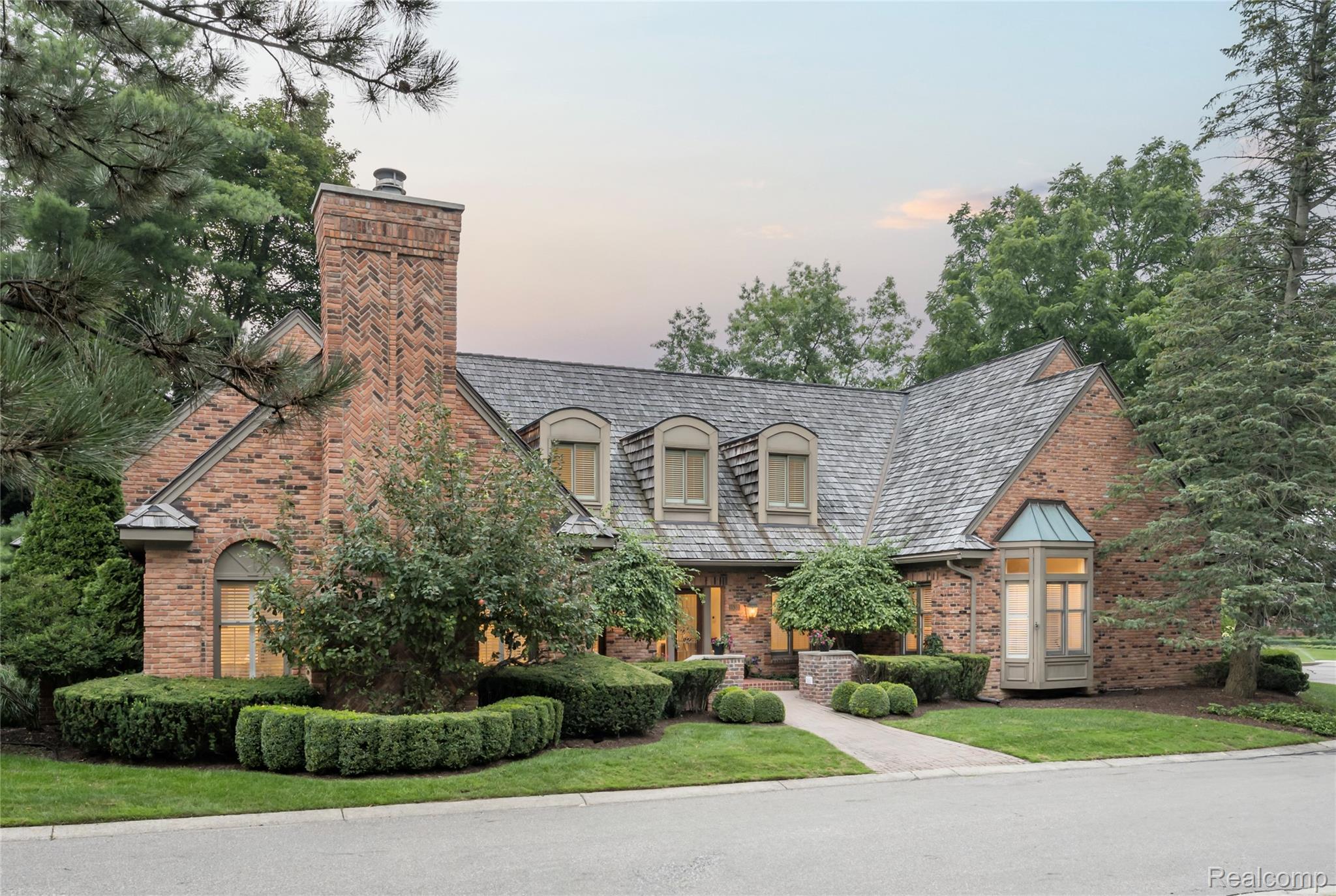 This is a front exterior view of a stately brick home with a prominent chimney and a gray shingle roof. The landscaping is meticulously maintained with trimmed hedges and mature trees, creating a sense of established elegance. The home features multiple windows with warm interior lighting visible, suggesting a cozy and inviting atmosphere.