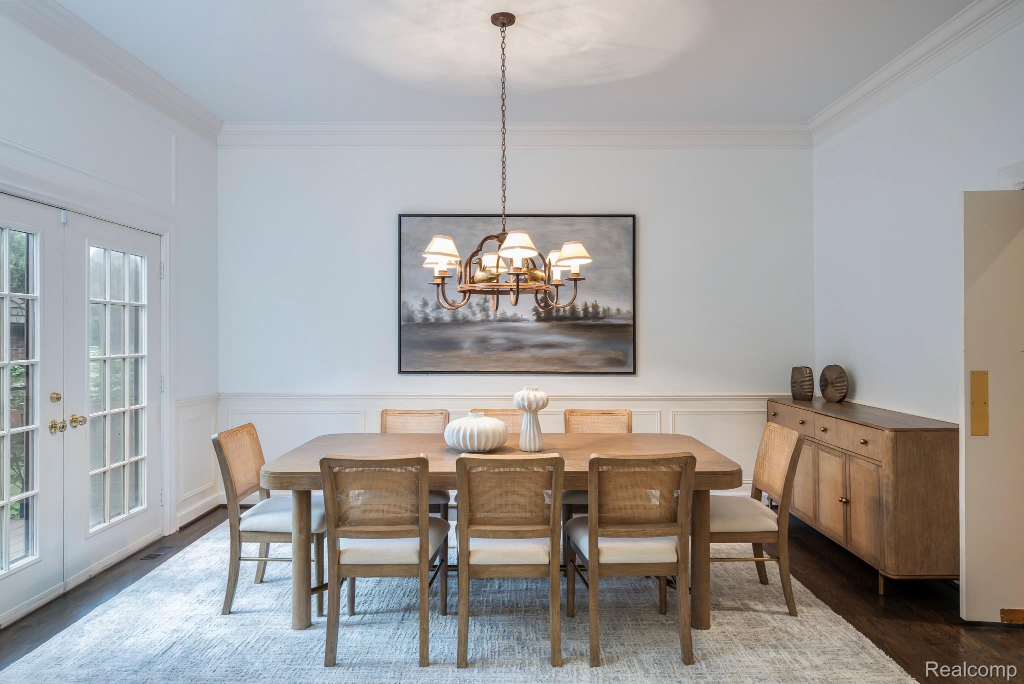 This is a well-lit dining room featuring a large wooden table with six chairs, a modern chandelier, and a framed landscape painting. A wooden sideboard sits against the wall, adding storage and style. The room is decorated in neutral tones, creating a warm and inviting atmosphere.