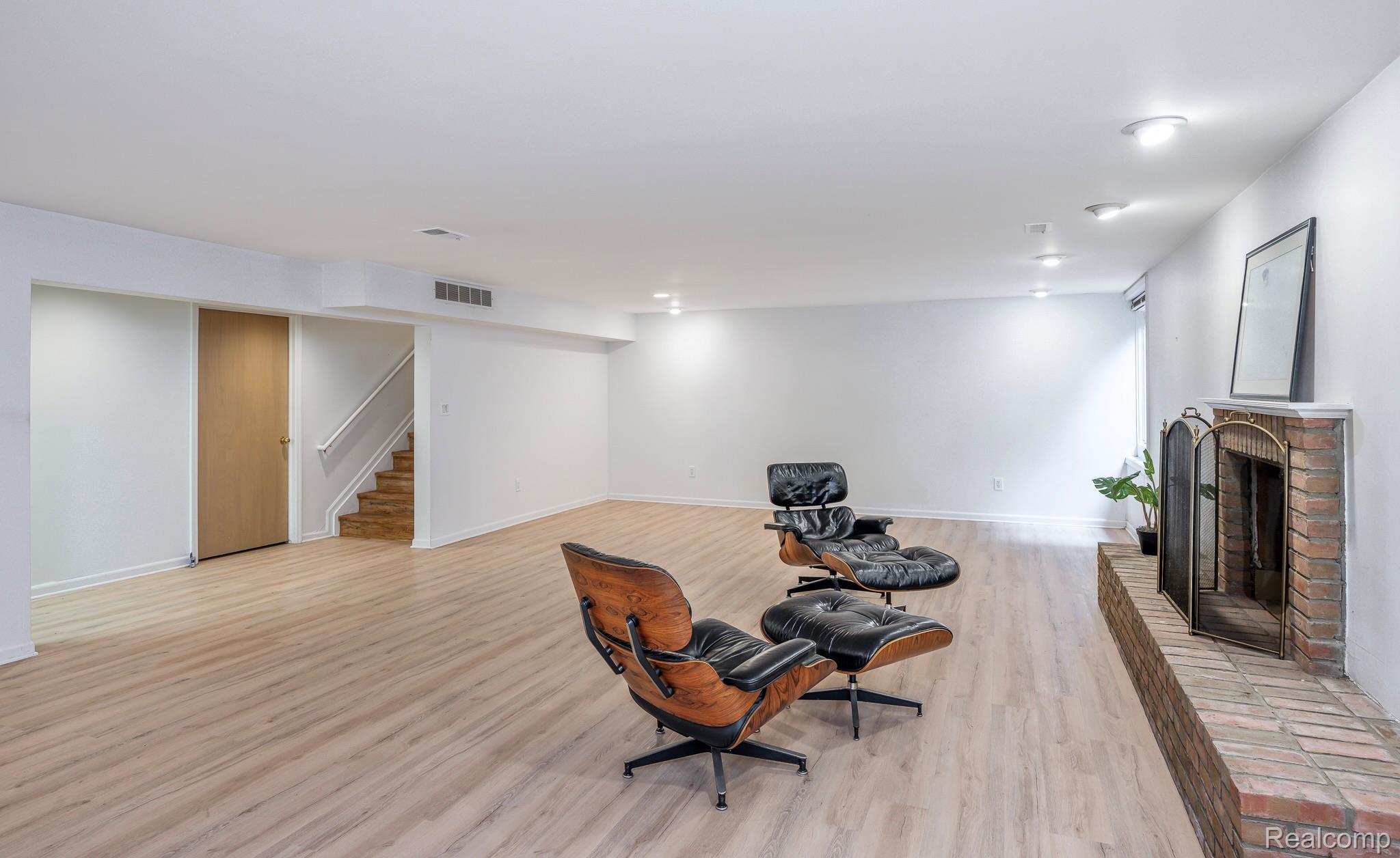 This is a spacious finished basement featuring light wood-look flooring, white walls, and recessed lighting. Two modern lounge chairs with ottomans are centrally placed, and a brick fireplace with a decorative screen adds a touch of warmth. A staircase and a closed door are visible on the left side of the image.