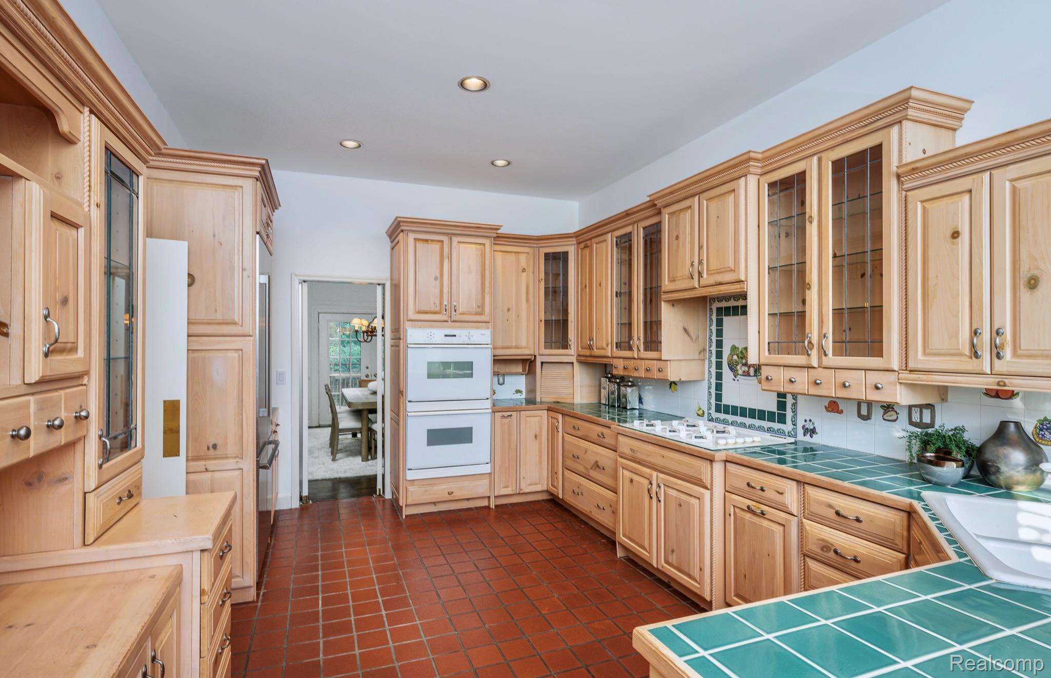 This is a kitchen with light wood cabinetry, including glass-fronted cabinets, and green tile countertops. The floor is tiled in a terracotta color, and there are built-in appliances such as a double oven. The kitchen has a warm, traditional style with a view into an adjacent dining area.