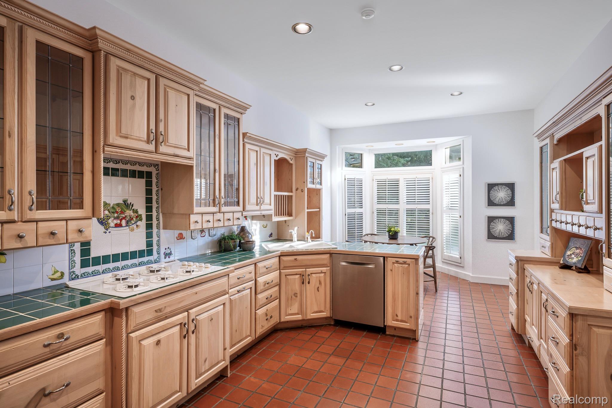 This is a kitchen with light wood cabinets, green tile countertops, and terracotta tile flooring. The kitchen features a built-in cooktop, a stainless steel dishwasher, and a bay window with white shutters. The overall style is traditional and cozy, with a focus on natural materials.