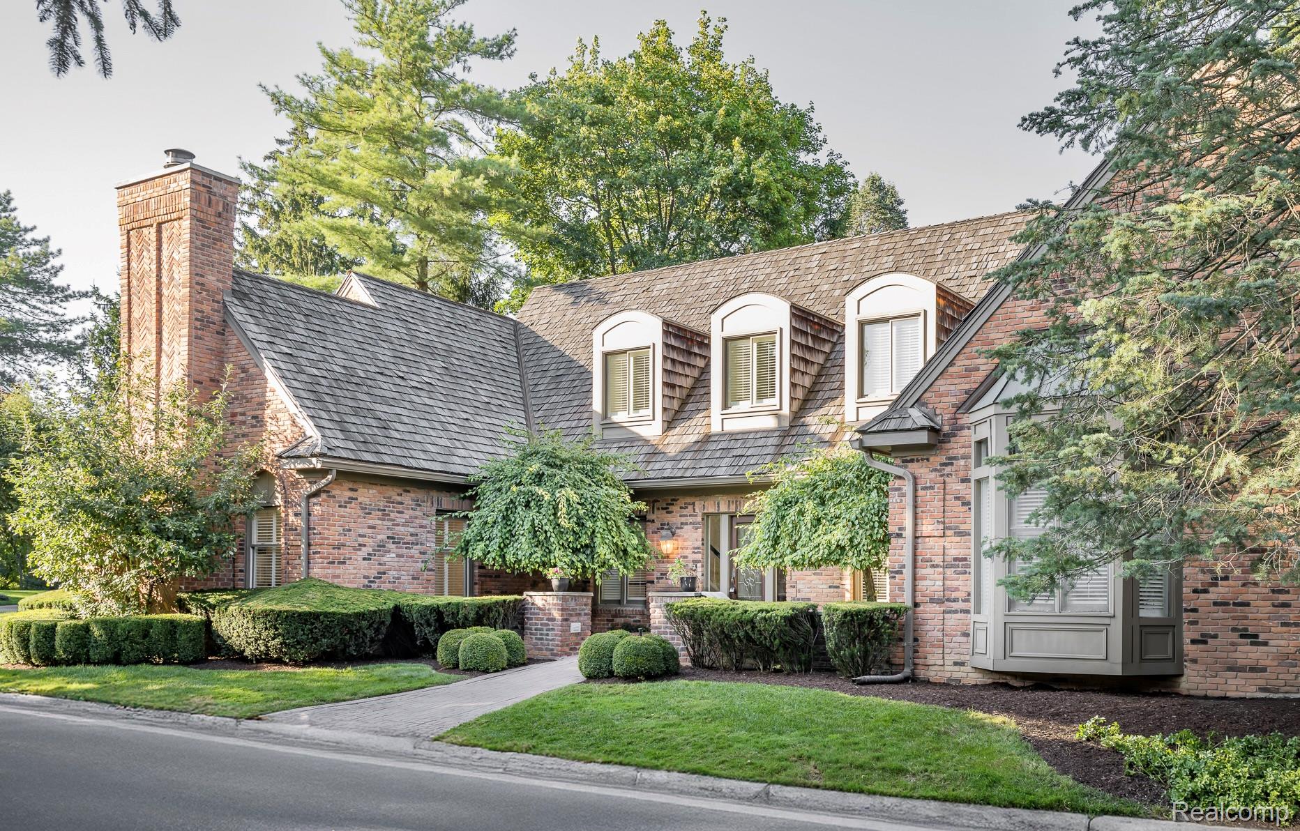 This is a front exterior view of a brick house with a complex roofline featuring dormer windows. The property is well-landscaped with manicured bushes and trees, and a paved walkway leads to the entrance. The overall impression is one of established elegance and curb appeal.