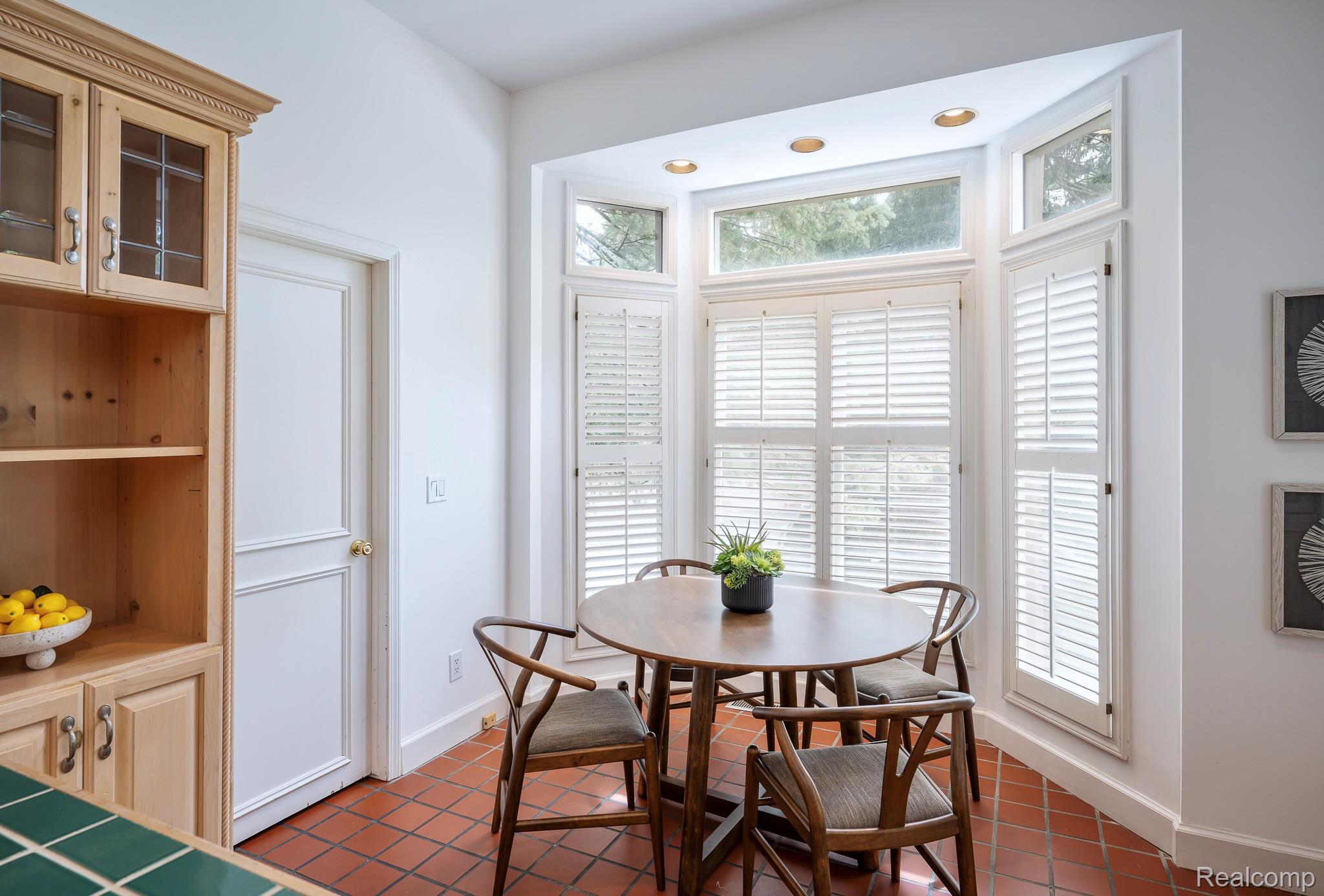This interior shot showcases a cozy dining area with a round wooden table surrounded by four chairs. A bay window with white shutters provides ample natural light, highlighting the terracotta tile flooring. A built-in wooden cabinet adds storage and character to the space, creating a warm and inviting atmosphere.