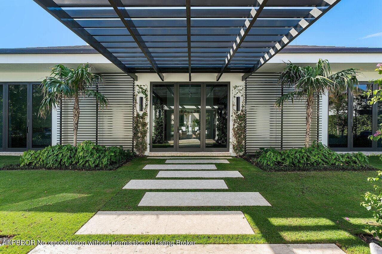 This image showcases a modern home's entryway, featuring a striking pergola above the entrance. A pathway of rectangular stone slabs leads to the glass front doors, flanked by symmetrical landscaping with palm trees and lush greenery. The architectural style is contemporary, emphasizing clean lines and a welcoming approach.