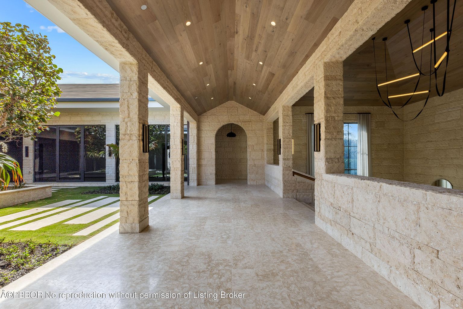 This image showcases a grand entryway featuring stone columns and walls, a wooden ceiling with recessed lighting, and a tiled floor. The design incorporates arched openings and modern light fixtures, creating a luxurious and inviting atmosphere. The perspective is from the entrance looking into the building.