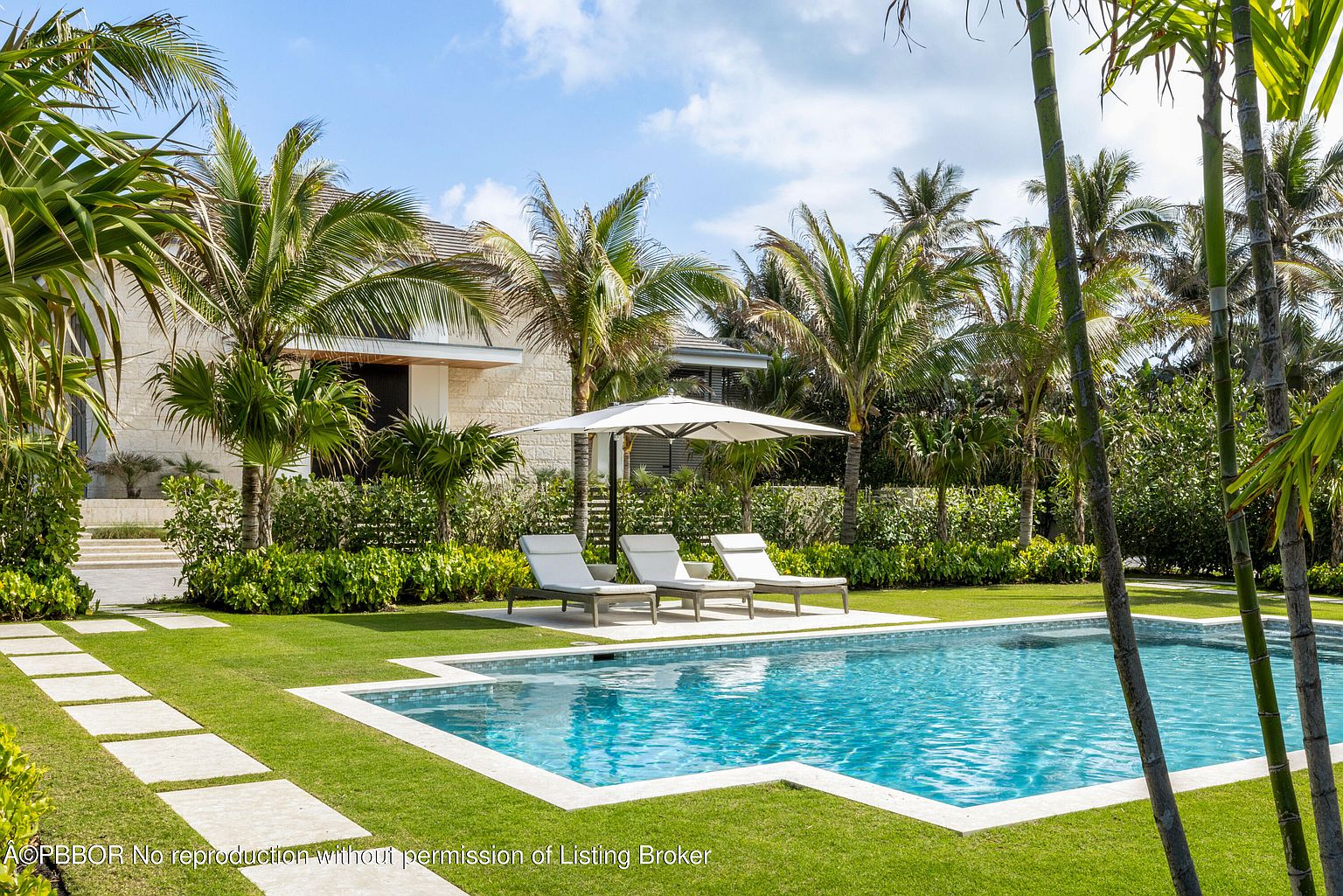 This image showcases a luxurious backyard pool area, complete with a pristine pool featuring a light-colored tile border. Three lounge chairs are neatly arranged under a large white umbrella, offering a relaxing space. The surrounding landscape includes lush green grass, palm trees, and manicured hedges, creating a private and serene oasis.