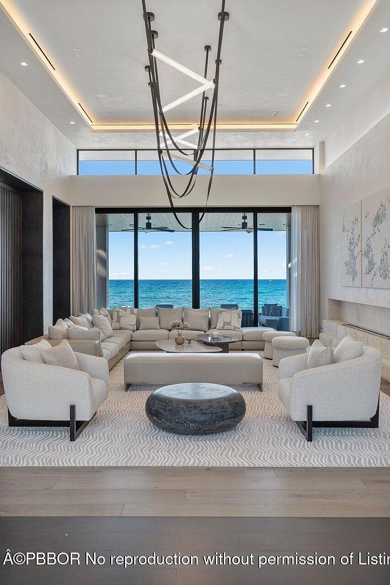 This is a luxurious living room with a large sectional sofa, two armchairs, and a unique coffee table arrangement. The room features floor-to-ceiling windows offering a stunning ocean view, complemented by a modern chandelier and neutral color palette. The overall impression is one of elegance and relaxation.