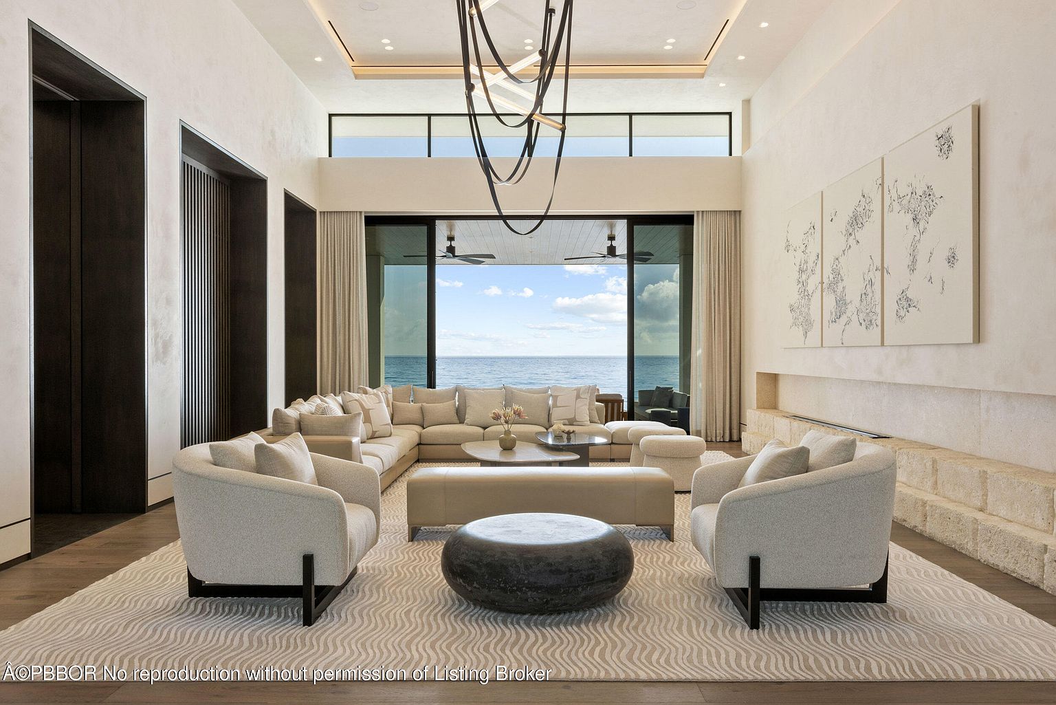 This is a luxurious living room featuring a large sectional sofa, two armchairs, and a unique coffee table arrangement. The room boasts floor-to-ceiling windows with ocean views, complemented by modern artwork and a sleek fireplace. The overall impression is one of sophisticated comfort and high-end design.