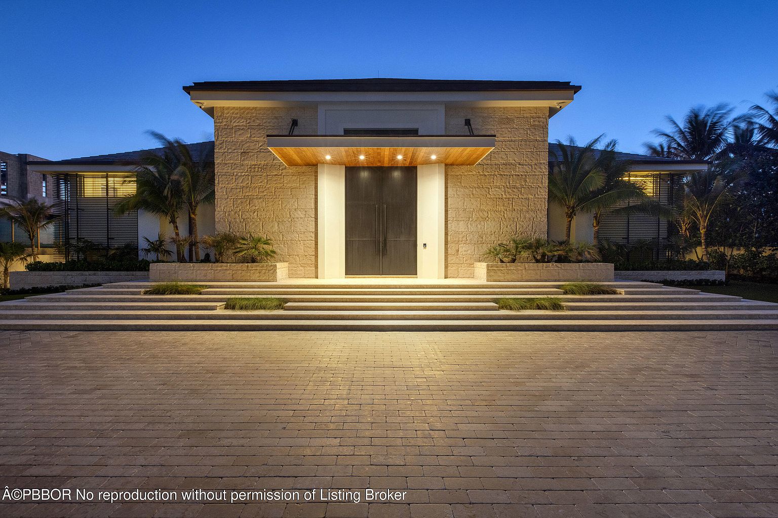This is a striking exterior shot of a home's entryway at dusk, featuring a grand entrance with a stone facade and a dark double door. The entrance is elevated with multiple tiers of steps, flanked by manicured greenery. The driveway is paved with stone bricks, and the overall impression is one of modern elegance and sophistication.