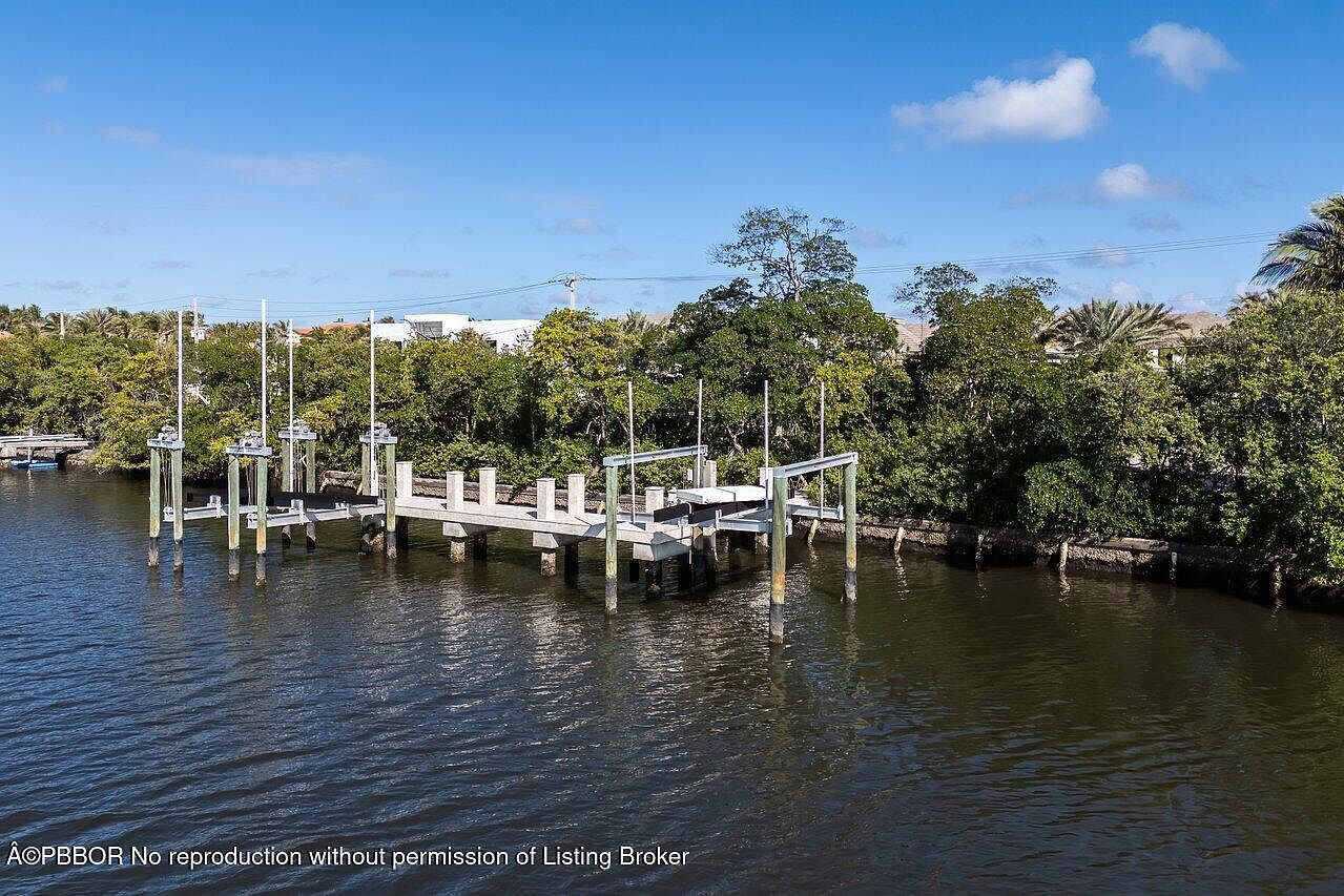 This aerial view showcases a private dock extending into calm, dark water, surrounded by lush green trees and vegetation. The dock features multiple boat lifts and a walkway, suggesting waterfront property with boating access. The sky is blue with scattered clouds, providing a serene and inviting atmosphere.