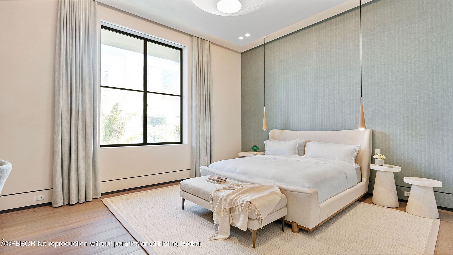 This is a bright and airy primary bedroom featuring a large window with natural light. The room is decorated in neutral tones with a light blue accent wall, creating a serene and relaxing atmosphere. The bed is neatly made with white linens, and there are stylish side tables and pendant lights on either side.