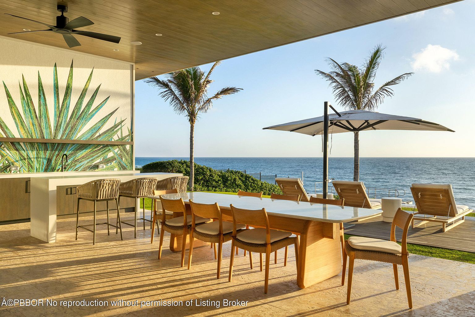 This image showcases a luxurious outdoor patio area with stunning ocean views. The space features a dining table with chairs, a bar area with stools, and lounge chairs under an umbrella, all designed for relaxation and entertainment. The presence of palm trees and the ocean backdrop create a tropical and serene atmosphere, perfect for upscale outdoor living.
