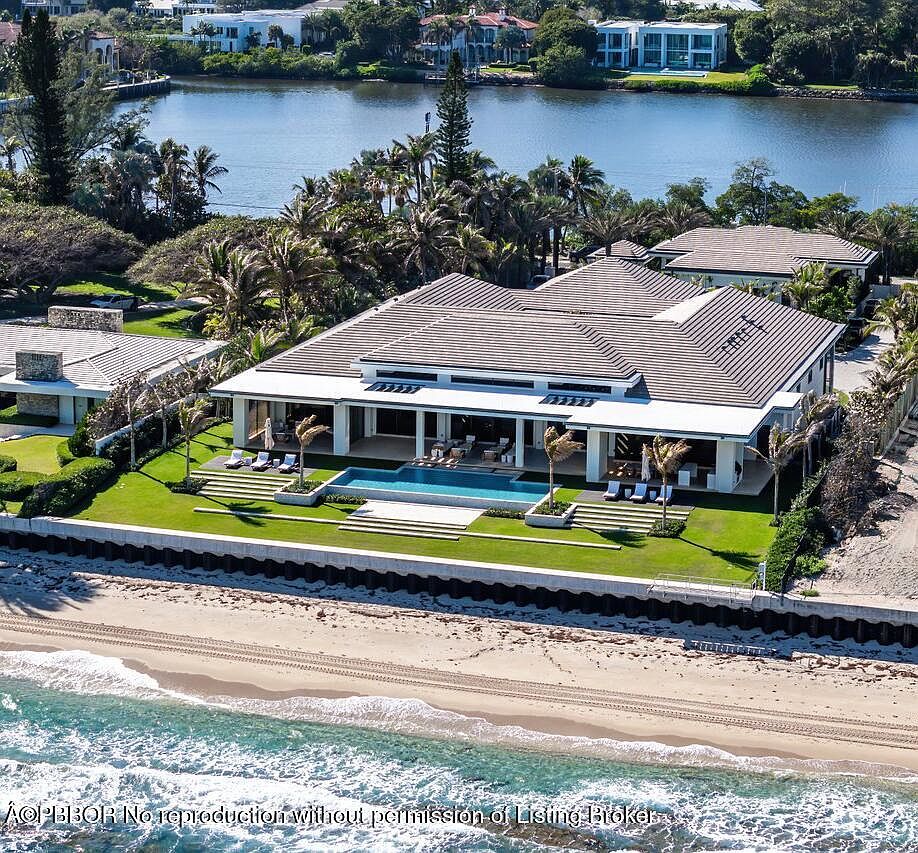 This aerial view showcases a luxurious beachfront property with a modern architectural design. The house features a large swimming pool, meticulously manicured lawns, and direct access to a sandy beach. The property is surrounded by lush tropical landscaping and offers stunning views of the water.