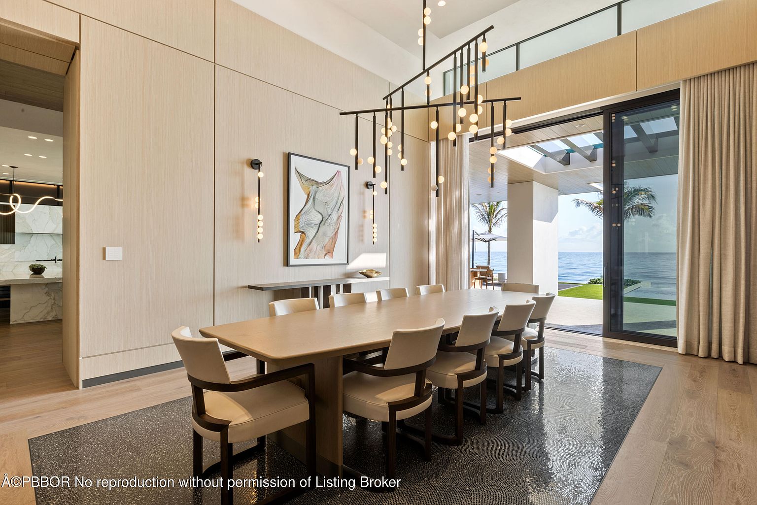 This is a bright and airy dining room featuring a long, light-colored wooden table surrounded by upholstered chairs with dark wood frames. A modern chandelier hangs above the table, and artwork adorns the walls. Large sliding glass doors offer a view of the ocean and provide access to an outdoor area, creating a seamless indoor-outdoor living experience.