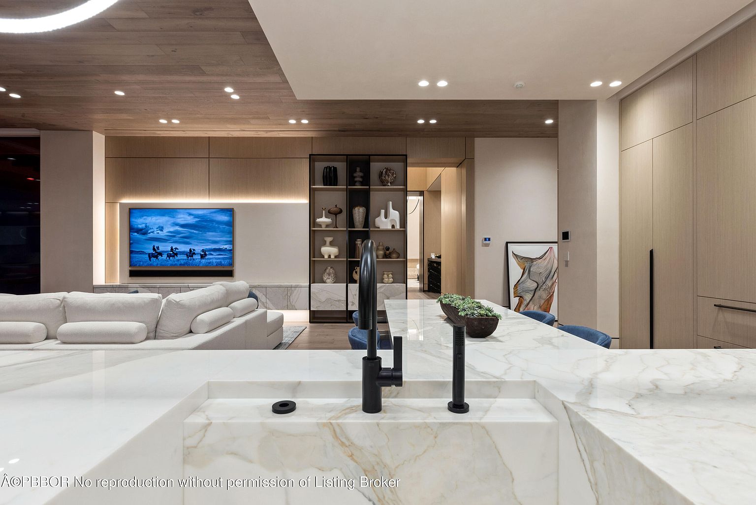 This interior shot showcases a modern kitchen and living space, featuring a large marble island with a black faucet as a focal point. The open-concept design includes a comfortable white sofa facing a large television, complemented by built-in shelving and wooden accents on the ceiling and cabinetry. The overall impression is one of luxury and contemporary elegance.