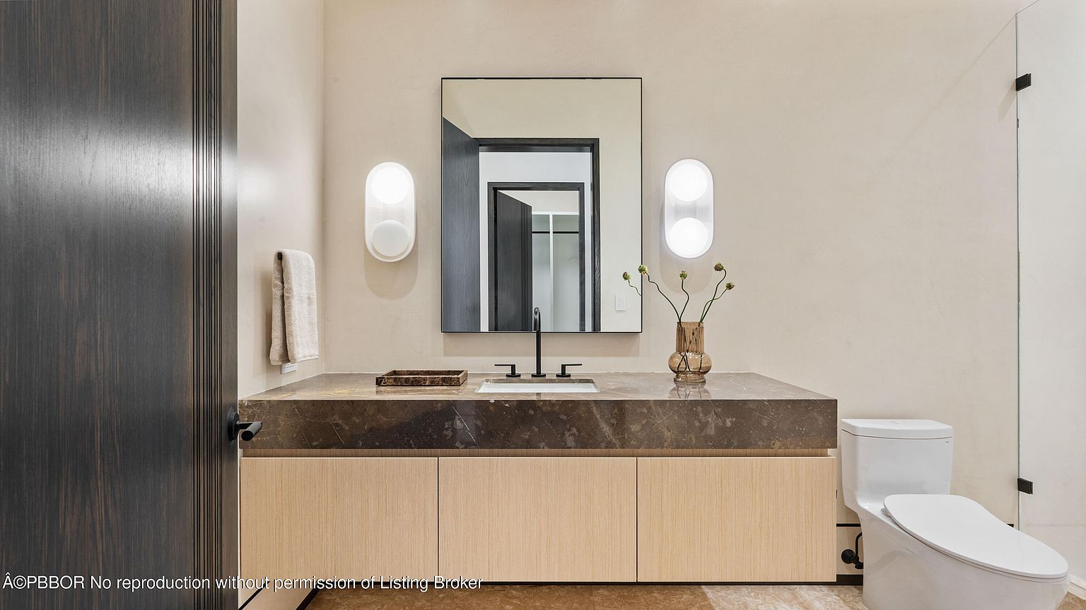 This is a modern bathroom featuring a floating vanity with a brown countertop and light wood cabinets. A rectangular mirror hangs above the sink, flanked by two vertical sconces. A toilet is visible to the right, and a dark wood door is on the left, creating a clean and contemporary aesthetic.