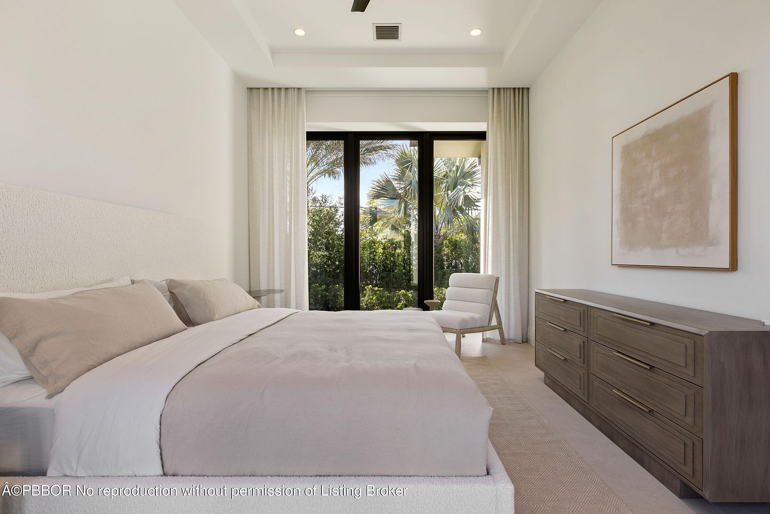 This is a bright and airy primary bedroom featuring a large bed with neutral-toned linens and a textured headboard. A modern dresser with sleek hardware sits against the wall, complemented by an abstract art piece. Large windows with sheer curtains offer a view of lush greenery, creating a serene and inviting atmosphere.