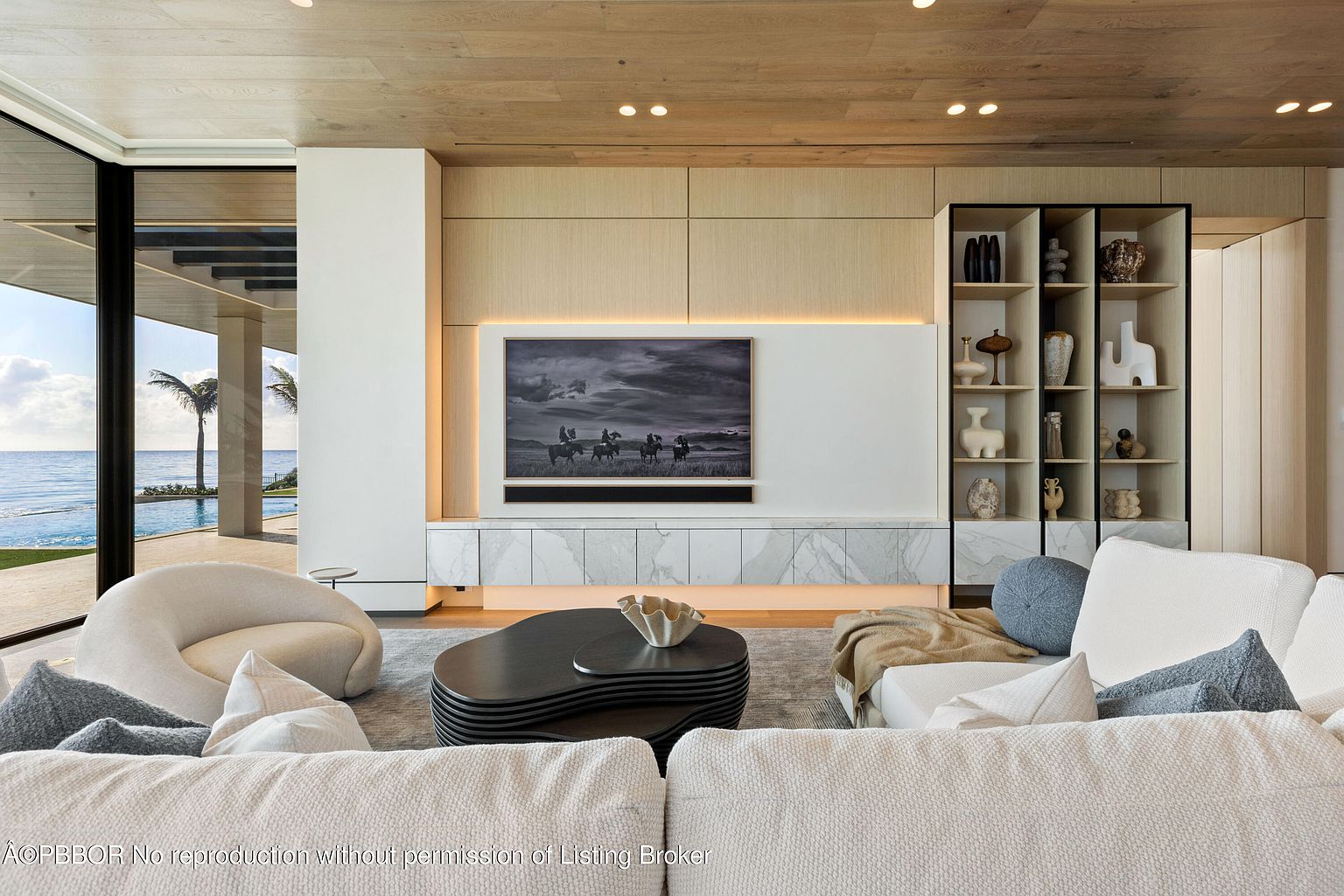 This is a luxurious living room featuring a modern design with a neutral color palette. The room includes a large sectional sofa, a unique coffee table, and a built-in entertainment center with a large screen TV and display shelves. Large windows offer a view of the ocean, enhancing the room's serene and upscale ambiance.