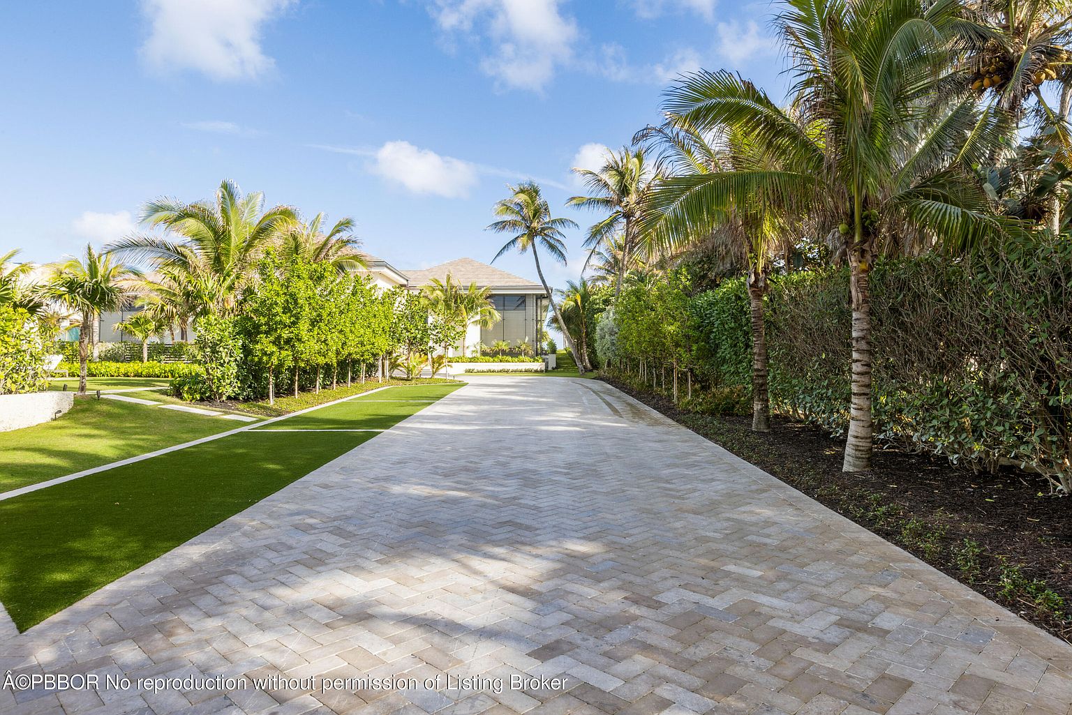 This image showcases the front exterior of a luxurious property, featuring a long, herringbone-patterned driveway leading to the house. Lush green lawns and meticulously maintained landscaping, including palm trees and manicured hedges, frame the driveway. The house itself is partially visible, suggesting a modern architectural style with clean lines and a neutral color palette, creating an inviting and upscale first impression.