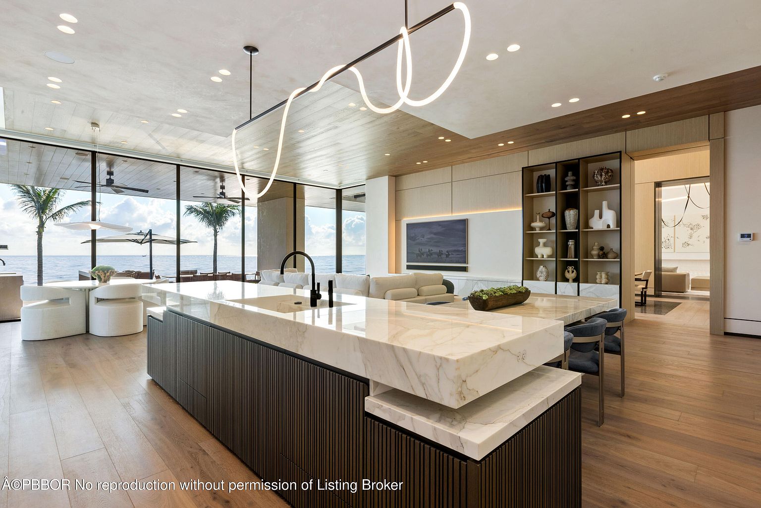 This interior shot showcases a modern, luxurious kitchen with a large island featuring a marble countertop and dark wood cabinetry. The open-concept design flows into a living area with ocean views visible through expansive windows. A unique, contemporary light fixture hangs above the island, adding a touch of elegance.