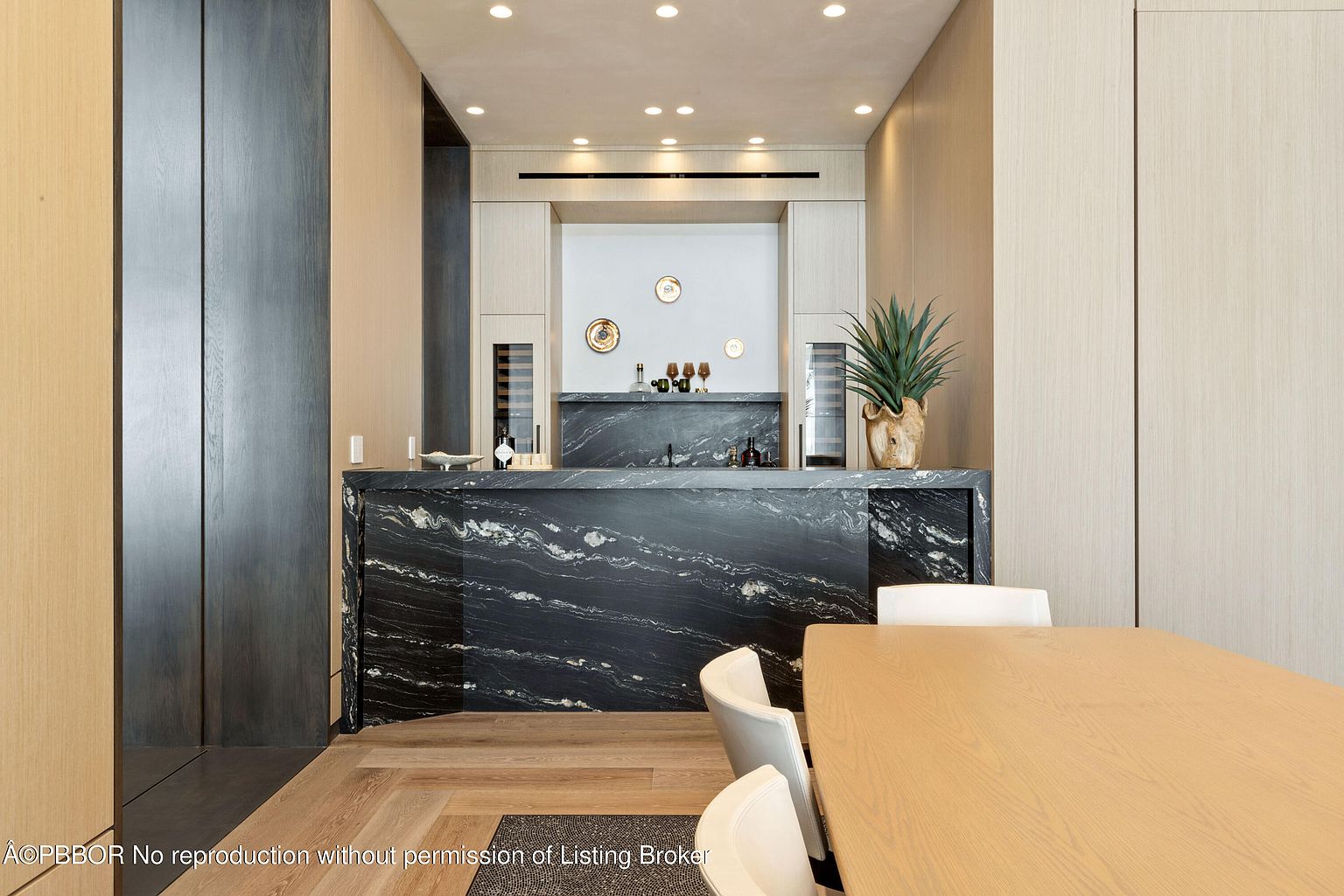 This interior shot showcases a dining area with a modern aesthetic. A dark marble countertop bar with built-in wine coolers anchors the space, complemented by light wood cabinetry and flooring. The dining table and chairs are contemporary, contributing to the room's sophisticated and minimalist design.