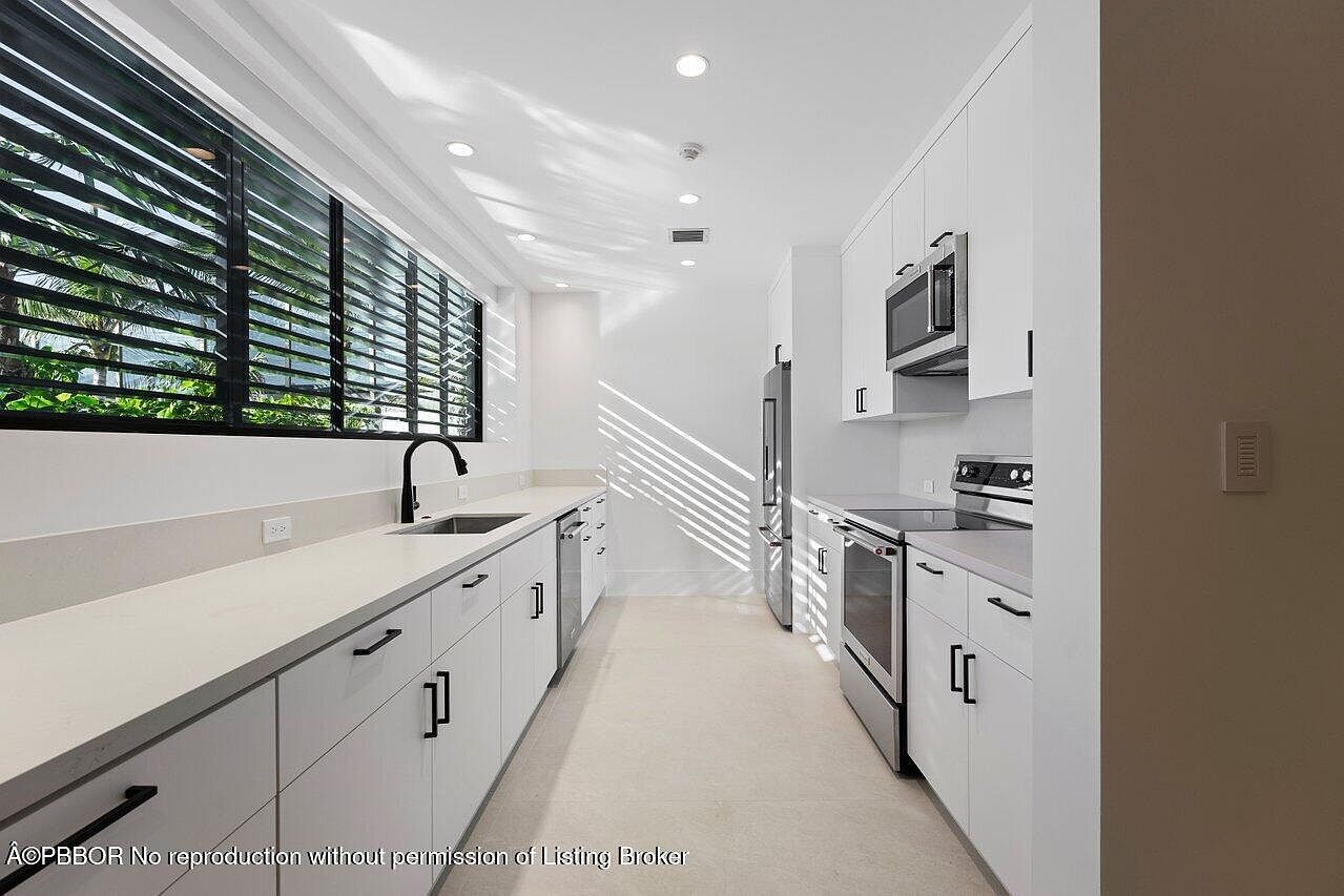 This is a well-lit, modern kitchen featuring white cabinetry with sleek, dark hardware. Stainless steel appliances, including a refrigerator, oven, and microwave, complement the clean aesthetic. A large window with black blinds provides natural light and a view to the outside, while the light-colored countertops and flooring enhance the bright and airy feel of the space.