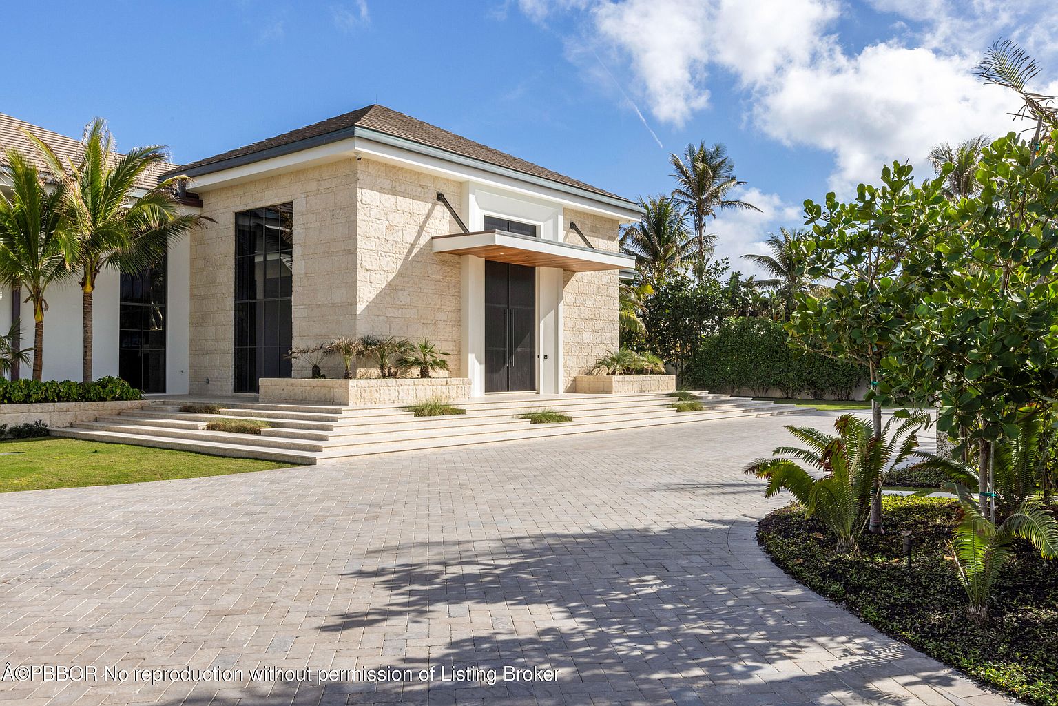 This is a front exterior shot of a modern, luxurious home, emphasizing the grand entryway. The house features a light stone facade, a covered entryway with a dark door, and multiple tiers of steps leading up to the entrance. The driveway is paved with light-colored stones, and the landscaping includes palm trees and manicured greenery, creating an inviting and upscale impression.