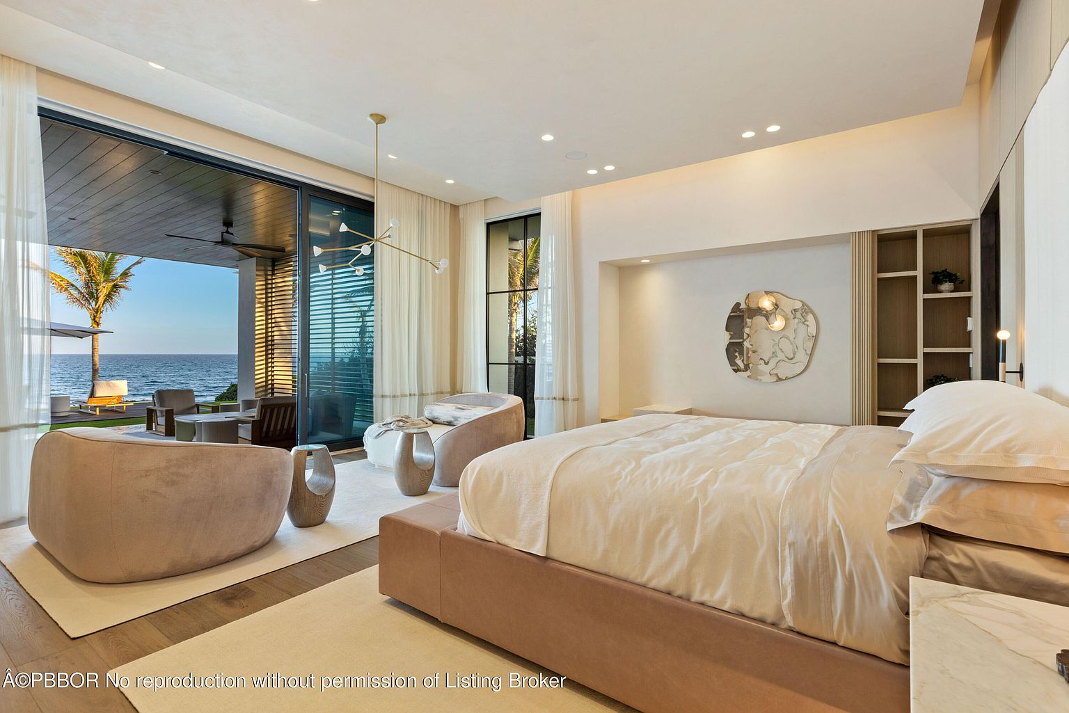 This is a luxurious primary bedroom featuring a king-size bed with plush bedding, modern lighting fixtures, and a serene ocean view through large sliding glass doors. The room is decorated in neutral tones with elegant furniture, creating a relaxing and sophisticated atmosphere. The perspective is from the corner of the room, showcasing the bed and the view.