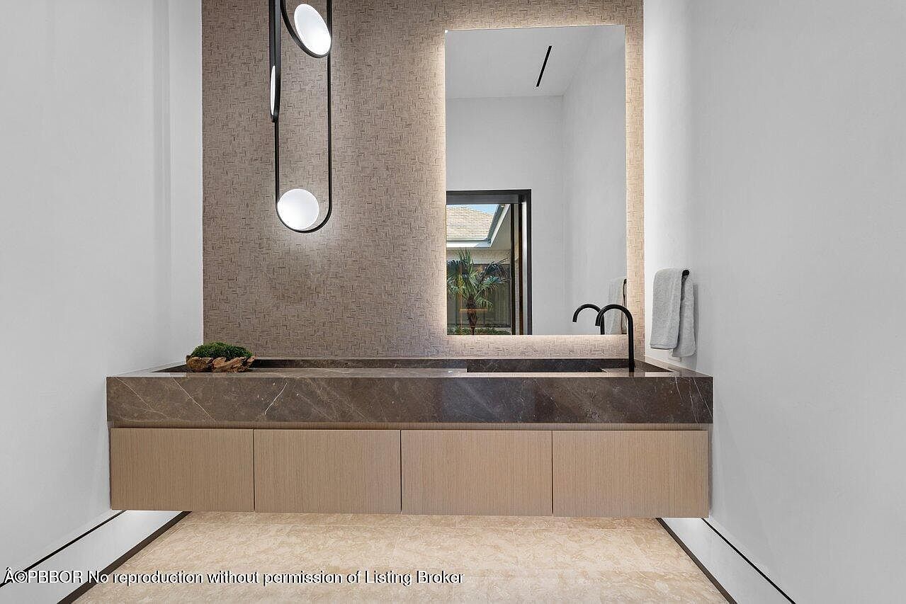 This is a modern bathroom featuring a floating vanity with a dark marble countertop and light wood cabinetry. A large, illuminated mirror hangs above the vanity, reflecting a glimpse of the outdoors. The space is lit by a contemporary pendant light fixture, and the overall design is sleek and minimalist.