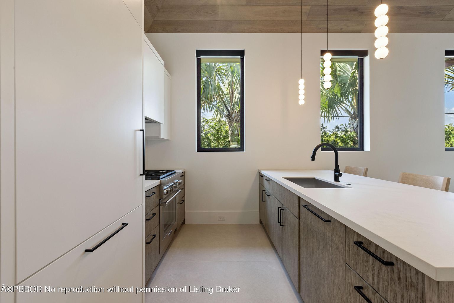 This is a modern kitchen featuring sleek white and wood-grain cabinetry with long, black handles. A stainless steel range sits next to the cabinets, and a large island with a white countertop dominates the center, featuring a black faucet and sink. Natural light streams in through two narrow windows, highlighting the clean lines and minimalist design.