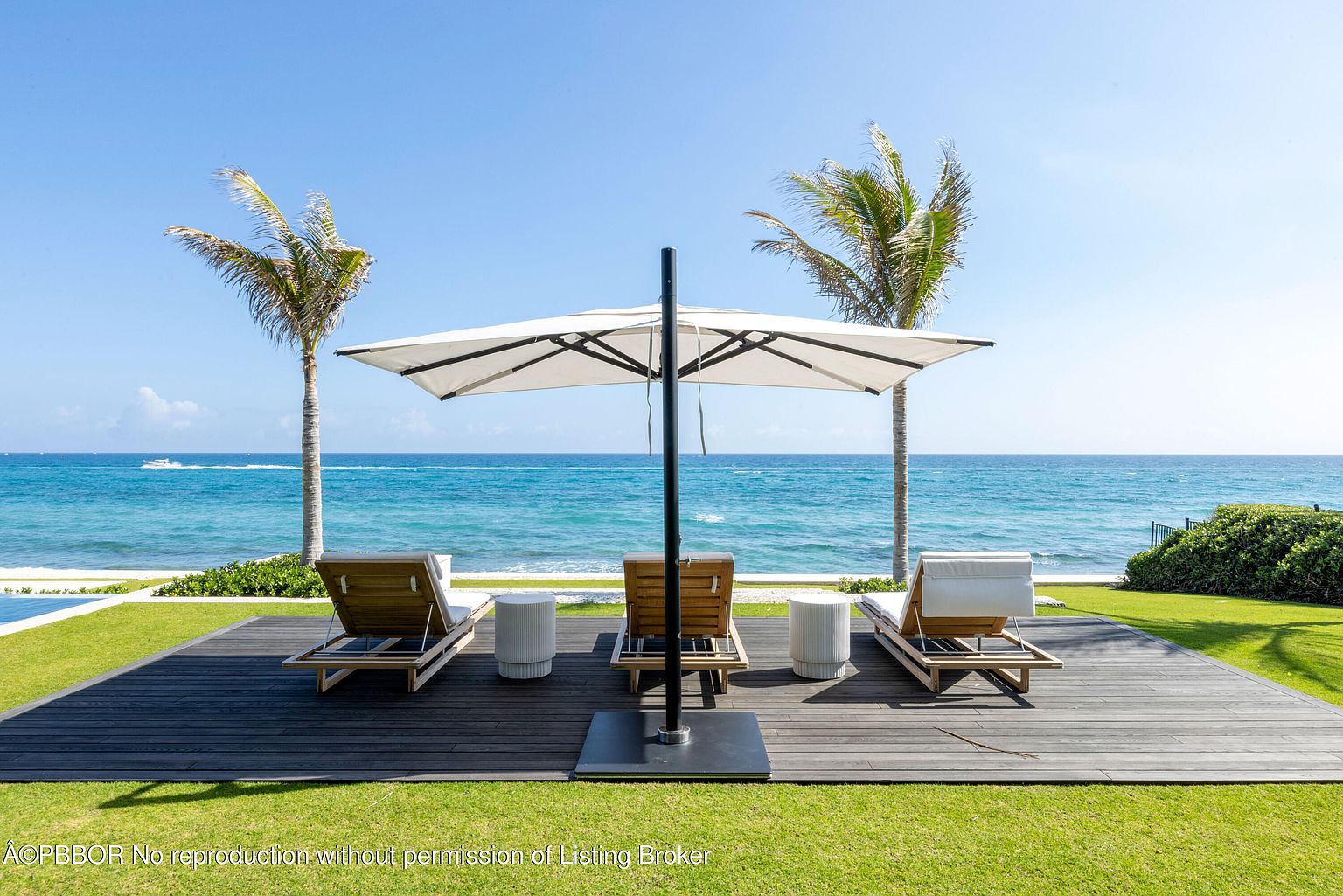 This exterior shot showcases a luxurious patio or deck area with a stunning ocean view. Three wooden lounge chairs are arranged under a large white umbrella, flanked by two palm trees, creating a relaxing and private outdoor space. The dark wooden deck contrasts beautifully with the bright green lawn and the turquoise ocean, enhancing the property's appeal.