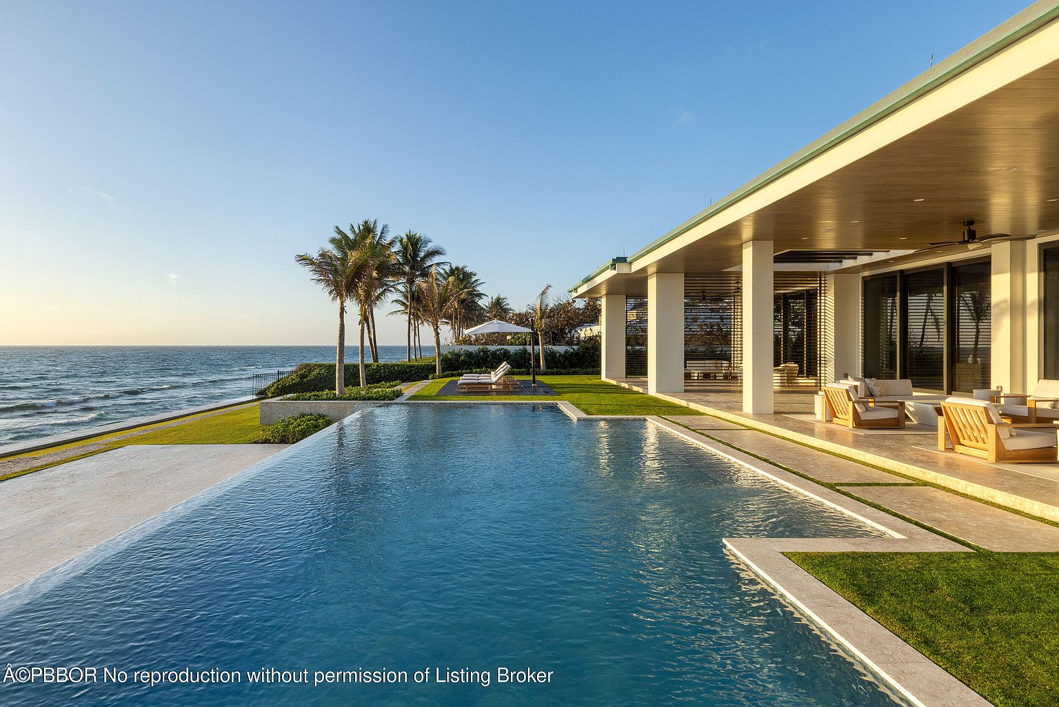 This image showcases a luxurious outdoor space featuring a stunning pool with clear blue water, seamlessly blending with the ocean view. The property boasts a modern architectural design with a covered patio area furnished with elegant seating. Palm trees line the horizon, adding to the tropical ambiance and highlighting the property's prime beachfront location.