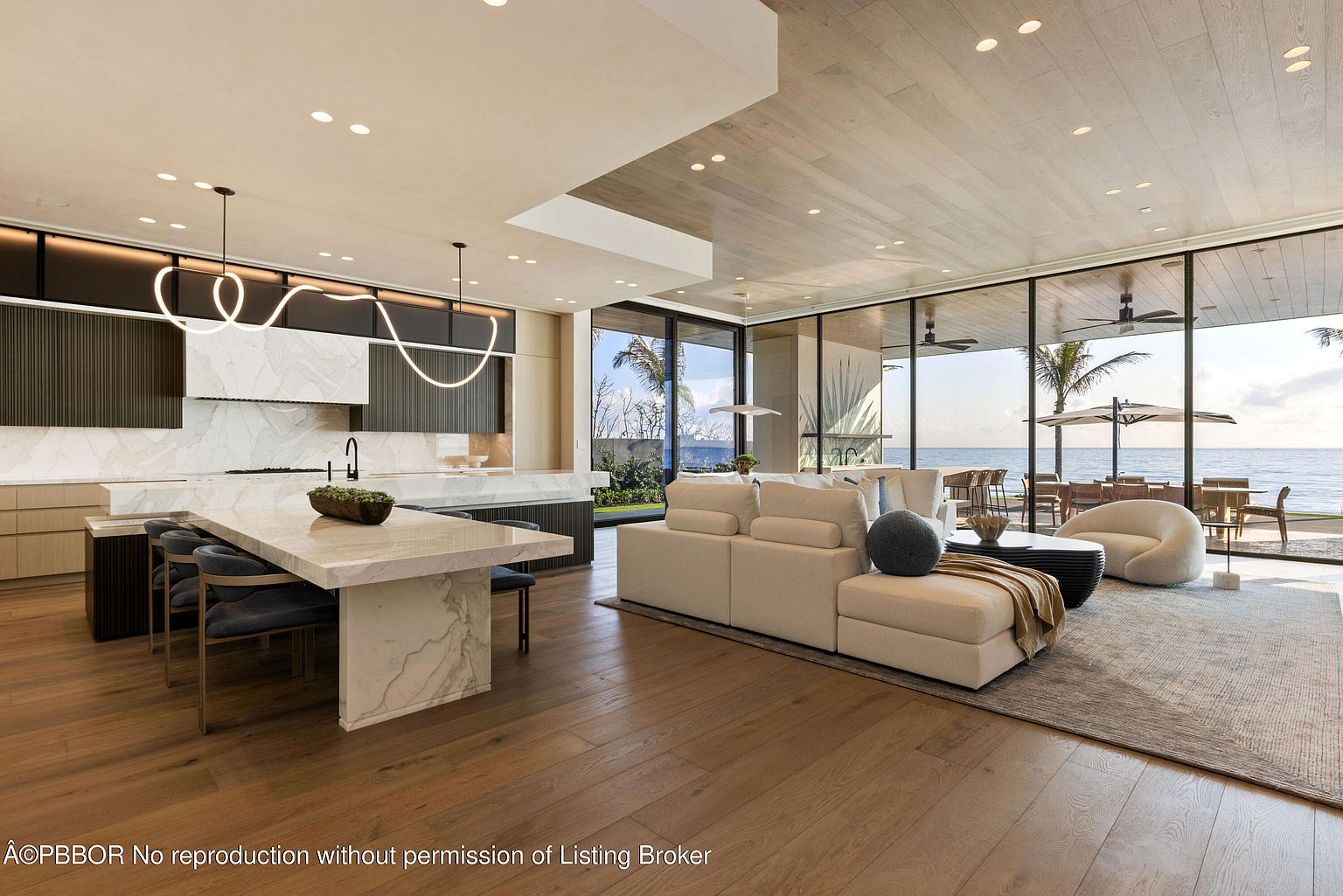 This is a wide-angle shot of a modern, open-concept kitchen and living area with floor-to-ceiling windows offering ocean views. The kitchen features a large marble island with seating, custom cabinetry, and unique pendant lighting. The living area includes a comfortable sofa and stylish decor, creating a seamless blend of luxury and functionality.