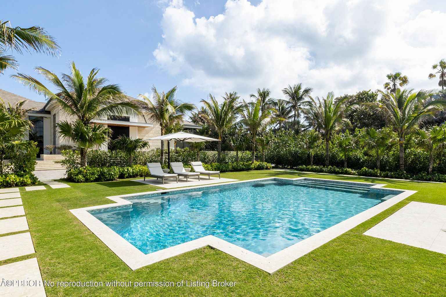 This image showcases a luxurious backyard pool area. The rectangular pool features clear blue water and is surrounded by a well-manicured green lawn, palm trees, and lounge chairs with an umbrella. The scene evokes a sense of relaxation and upscale outdoor living, perfect for entertaining or enjoying a private oasis.