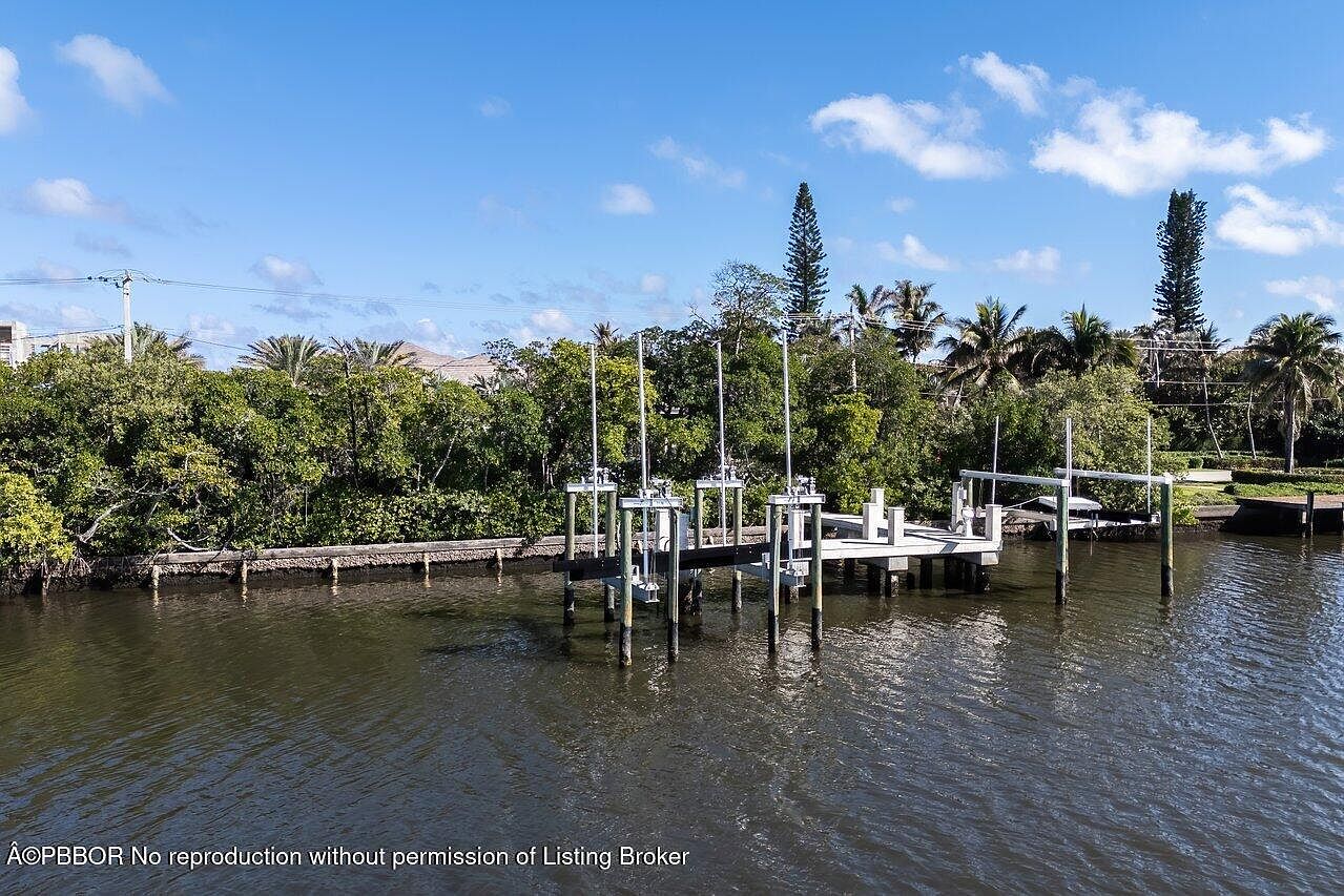 This image showcases the rear of a waterfront property, featuring a private dock with boat lifts. Lush greenery lines the shoreline, providing a natural backdrop. The water is calm, reflecting the clear blue sky with scattered clouds, creating a serene and inviting atmosphere.